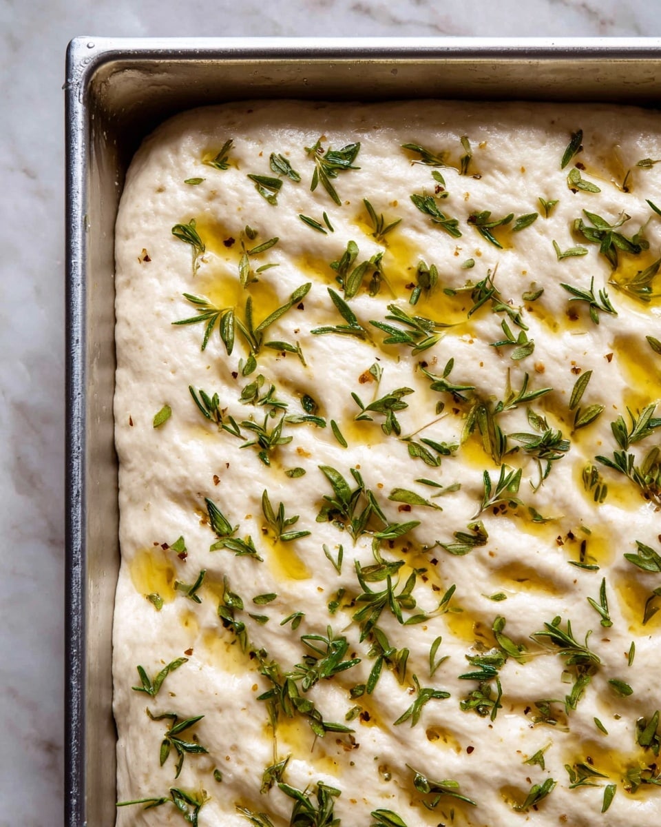 The image shows a close-up view of a thick, pale dough spread in a metal baking pan. The dough has visible dimples and a soft, slightly bubbly texture. It is topped with scattered fresh green herbs and drizzles of golden olive oil that add shiny spots to the surface. The background is a white marbled texture. photo taken with an iphone --ar 4:5 --v 7