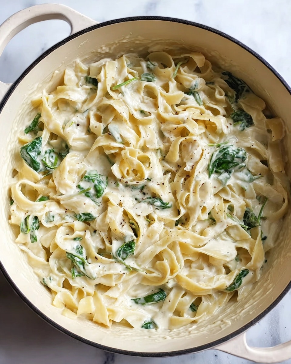 A white pot filled with soft, wide pasta noodles coated in a creamy white sauce. Mixed in between the pasta are fresh green spinach leaves that add a splash of color. The sauce looks smooth and thick, sticking well to the noodles. The pot is set on a white marbled surface. photo taken with an iphone --ar 4:5 --v 7
