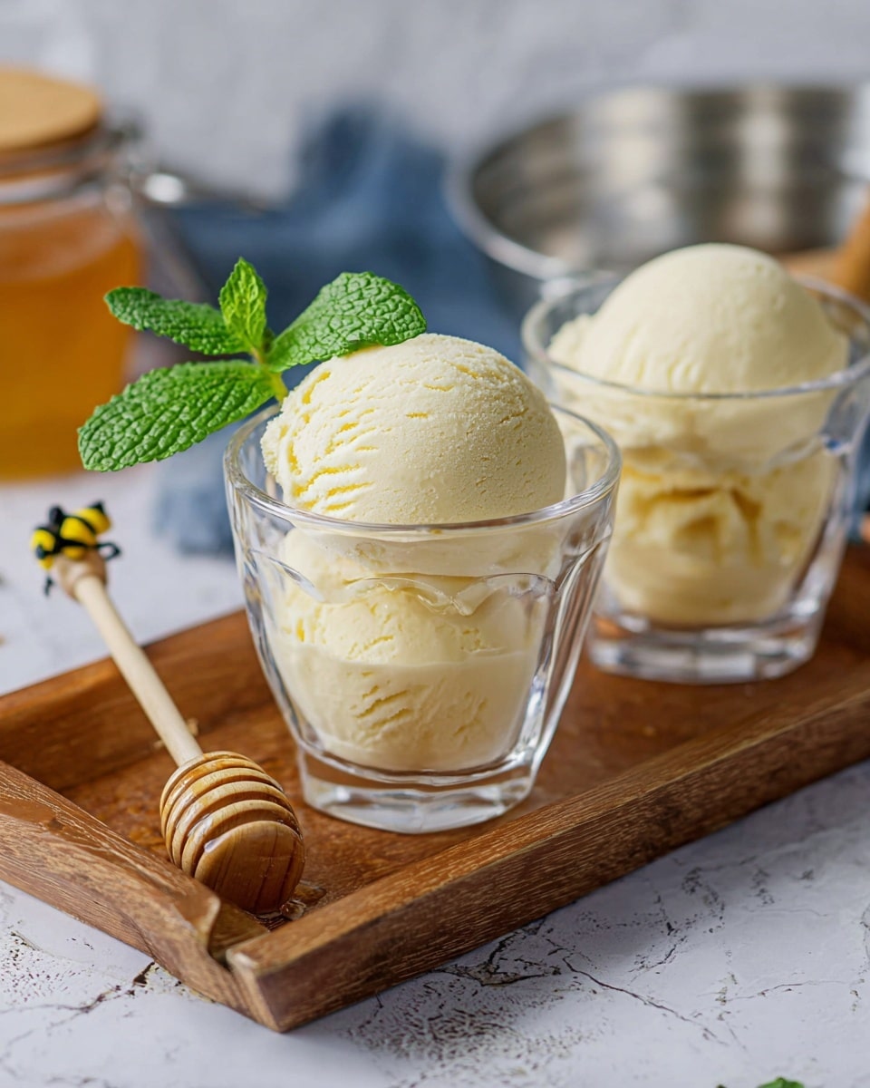 Two clear glass cups each hold one scoop of pale yellow ice cream, with a fresh green mint leaf tucked behind the scoop in the front cup. The cups are placed on a wooden tray that sits on a white marbled surface. Next to the front cup is a small wooden honey dipper with a tiny yellow and black bee decoration on its handle. In the background, part of a silver container and a honey jar with a wooden lid are visible. Photo taken with an iphone --ar 4:5 --v 7