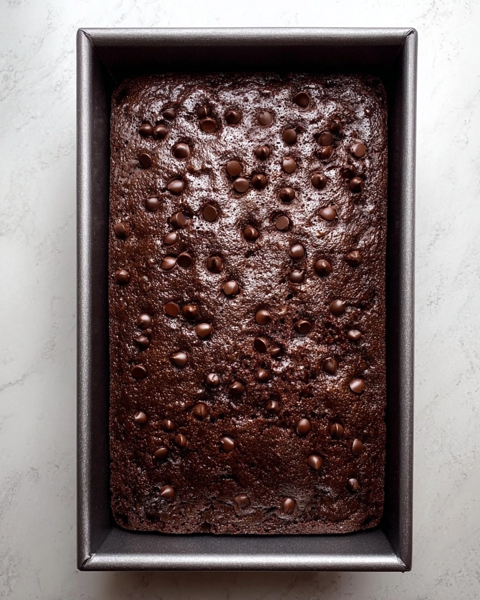 A rectangular dark brown chocolate cake with a slightly glossy surface covered in scattered chocolate chips, sitting inside a metal baking pan. The cake has a textured top with small bumps and cracks, indicating it is baked but still moist. The pan is placed on a white marbled surface. photo taken with an iphone --ar 4:5 --v 7