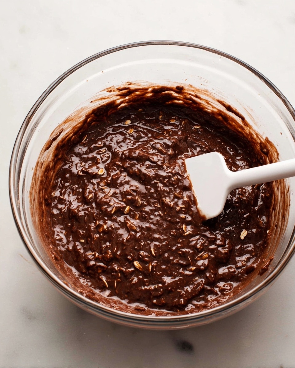 A clear glass mixing bowl is filled with thick, dark brown chocolate batter mixed with oats that give a rough texture throughout. The batter looks moist and slightly shiny, with visible oat flakes scattered evenly inside. A white spatula with a smooth handle is resting inside the bowl, partly covered with the chocolate oat mixture. The bowl sits on a white marbled surface. photo taken with an iphone --ar 4:5 --v 7