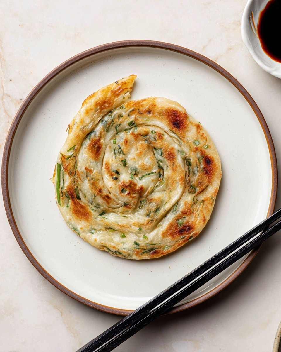 A single round spiral of flatbread with three visible layers is placed at the center of a white plate with a thin brown rim. The outer layer is golden brown and crispy, with small darker spots. Inside layers show pale dough interspersed with green bits of scallions or herbs, creating a textured look with some layers slightly separated. Beside the plate, a pair of black chopsticks rests diagonally on the white marbled surface. A small white bowl with a dark soy sauce is partly visible in the top right corner. The photo taken with an iphone --ar 4:5 --v 7