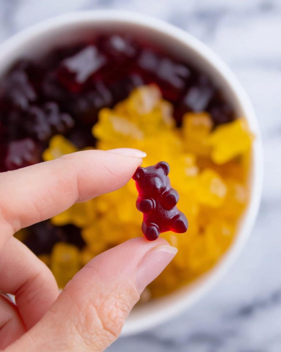 A close-up view shows a woman's hand holding a small, dark red gummy bear candy between thumb and finger. In the blurred background, there is a white bowl filled with gummy bears in two colors: dark red and bright yellow, mixed together. The bowl sits on a white marbled surface, and the photo focuses sharply on the gummy bear between the woman's fingers while the rest is softly blurred. photo taken with an iphone --ar 4:5 --v 7