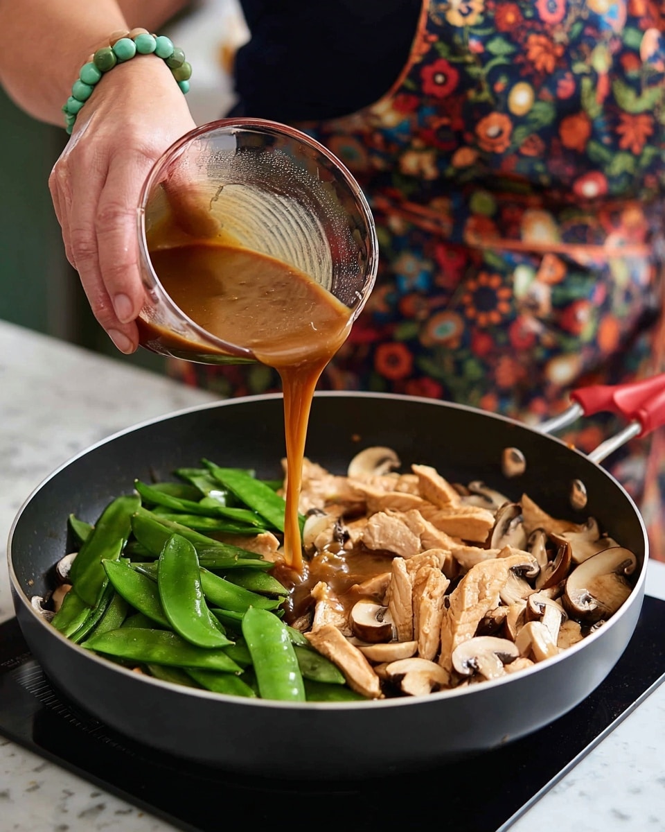 A black pan on a white marbled surface holds a stir-fry mix with three main layers: the bottom layer appears as light brown cooked chicken slices, the middle layer has bright green snap peas with a smooth texture, and the top layer features thinly sliced light beige mushrooms scattered evenly. A woman's hand wearing a jade green bracelet pours a smooth, medium brown sauce from a clear glass measuring cup into the pan. The person is wearing a dark shirt and a colorful apron with various patterns. Photo taken with an iphone --ar 4:5 --v 7