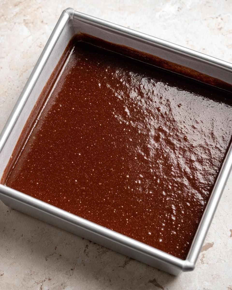 The image shows a square silver baking tray filled with a smooth, thick layer of shiny dark brown chocolate batter, evenly spread to the edges. The surface of the batter has a slightly glossy and wet texture with small bubbles scattered across. The tray is placed on a white marbled textured surface. Photo taken with an iphone --ar 4:5 --v 7