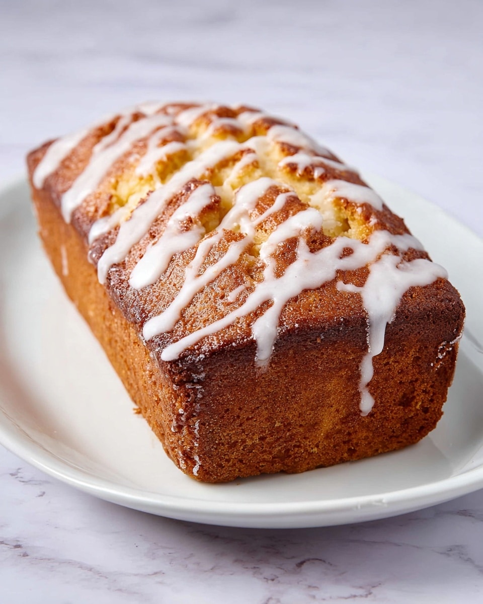 A golden brown loaf cake with a slightly darker crust sits on a white round plate. The cake has a textured surface with cracks running across the top, showing a soft, lighter yellow inside. White icing is drizzled in thin, uneven lines over the top, adding a shiny, smooth contrast to the rough cake texture. The plate rests on a white marbled surface. photo taken with an iphone --ar 4:5 --v 7