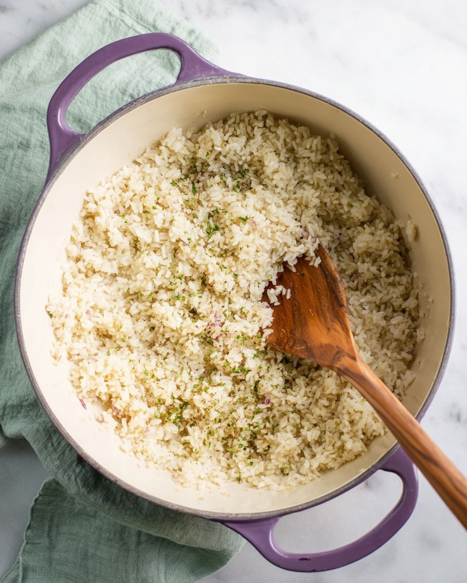 The image shows a purple-handled large white pot with a cream inside where small grains of cooked rice mixed with small bits of onions and herbs cover the bottom in a single thick layer. A wooden spoon with a long handle is placed inside the pot, partially submerged in the rice, with the spoon's flat end resting on the rice. The pot is sitting on a white marbled surface and a pale green cloth is seen in the upper left corner. photo taken with an iphone --ar 4:5 --v 7