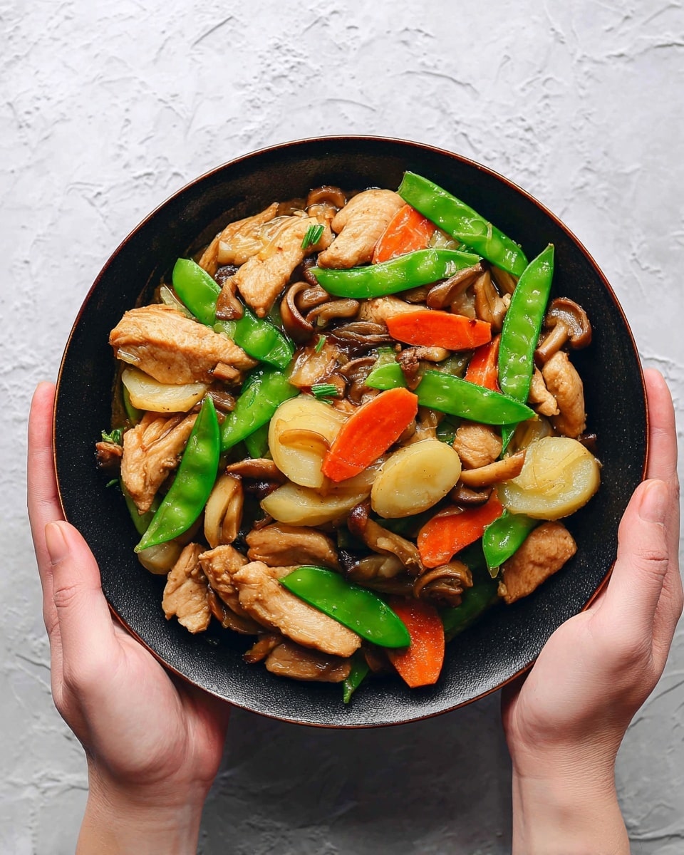 A dark bowl filled with cooked light brown chicken pieces layered with bright green snap peas, orange carrot slices, pale yellow water chestnuts, and light brown mushroom slices, all mixed together with a slight shiny sauce. The bowl is held by a woman's hand on the right and another woman's hand on the bottom, against a white marbled textured surface. photo taken with an iphone --ar 4:5 --v 7