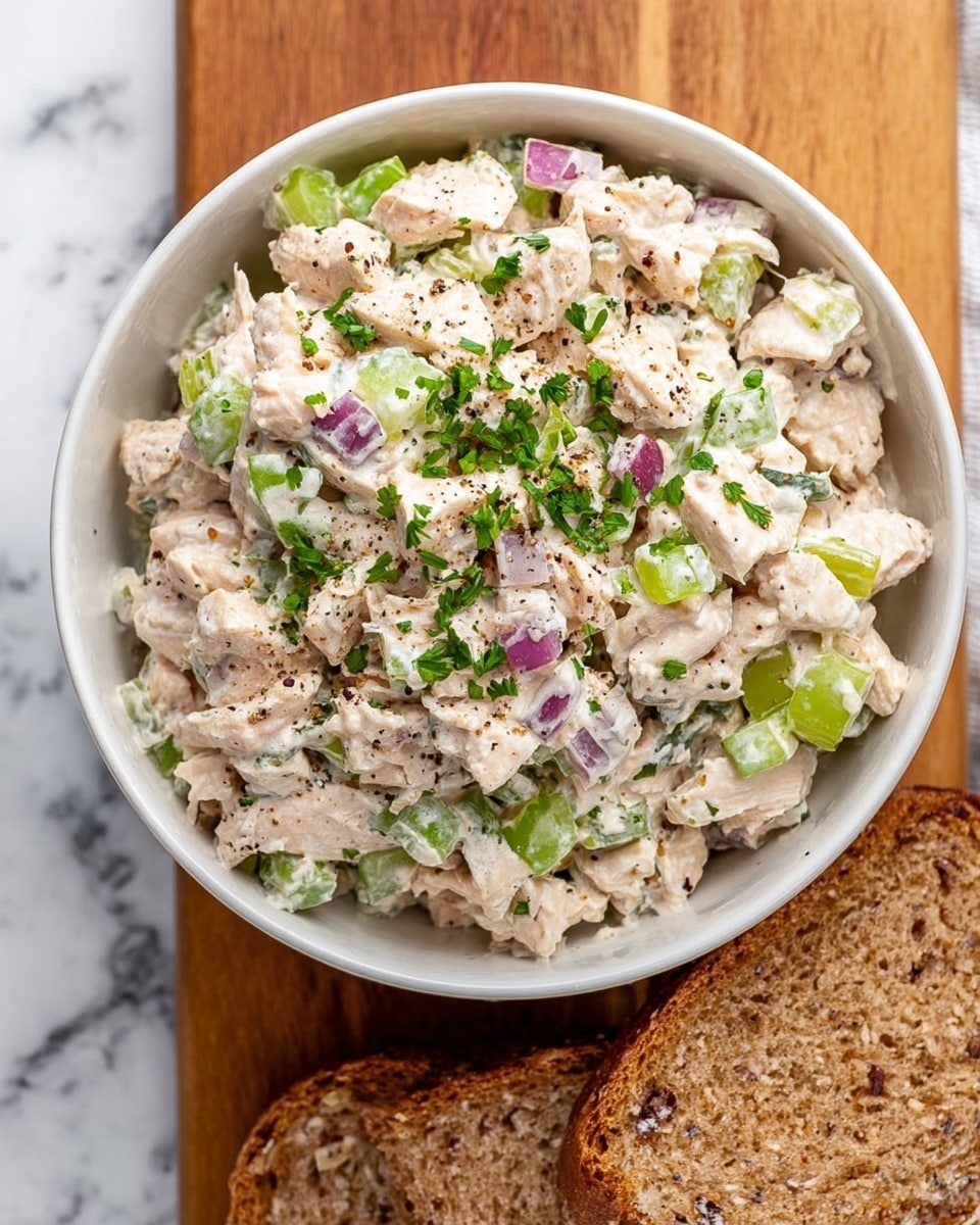A white bowl filled with a mixed chicken salad that has a creamy texture and is mostly pale beige with small chunks of light brown chicken. The salad is dotted with bright green celery pieces, purple onion bits, and finely chopped green herbs scattered on top and throughout, with a light sprinkling of black pepper. The bowl sits on a wooden board with two slices of multigrain bread at the bottom right edge. The background is a white marbled surface. Photo taken with an iphone --ar 4:5 --v 7