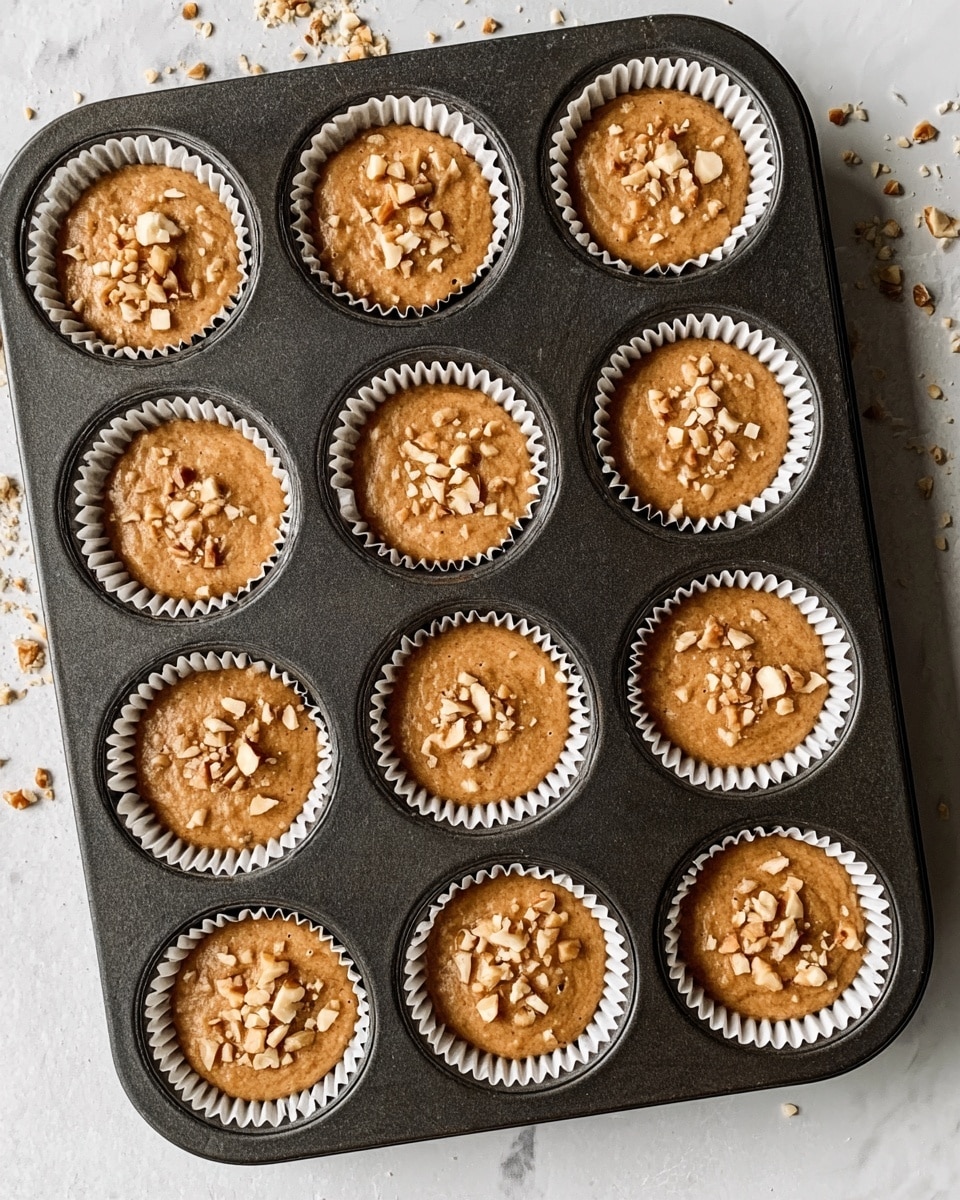 A dark metal muffin tray holds twelve white cupcake liners filled with smooth, light brown batter. Each cupcake is topped with small pieces of chopped nuts scattered evenly across the surface. The tray is placed on a white marbled surface with some nut crumbs around it, adding a casual, homemade feel. The texture of the batter looks slightly thick but even. photo taken with an iphone --ar 4:5 --v 7