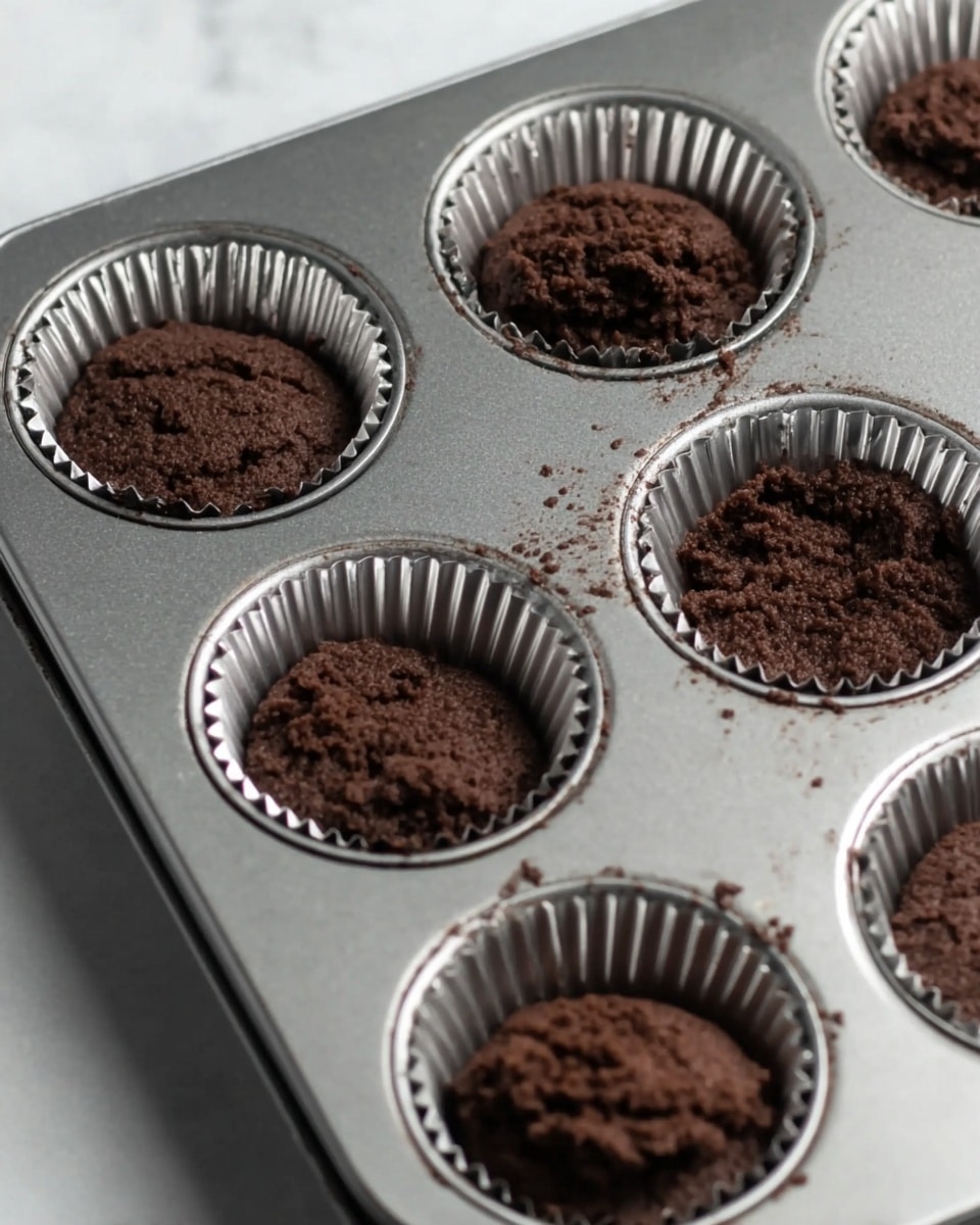 A dark brown soft chocolate batter is scooped inside shiny silver cupcake liners arranged in a metal muffin tray, filling each liner almost halfway. The batter looks thick and crumbly with a rich texture, resting inside six cups of the tray. The tray sits on a white marbled surface. Photo taken with an iphone --ar 4:5 --v 7