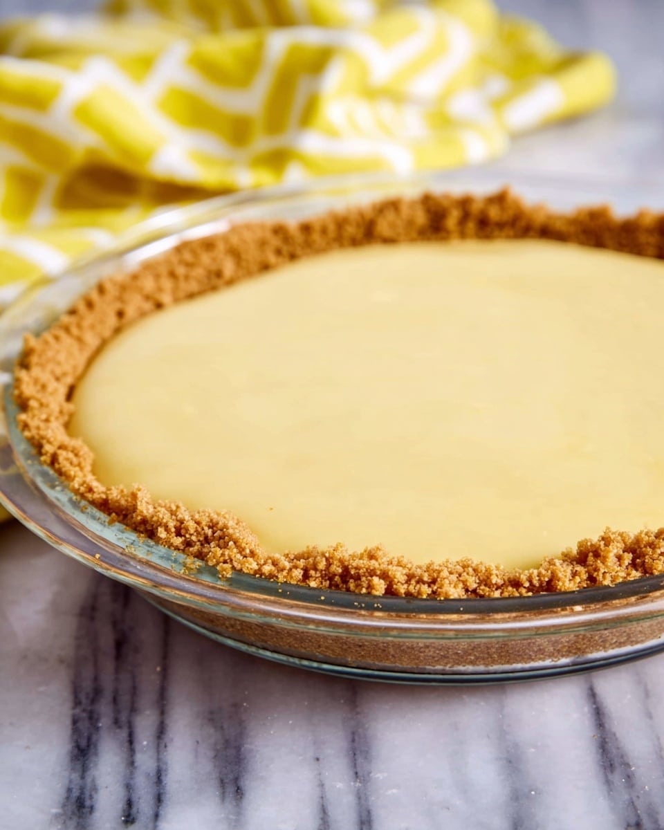 A clear glass pie dish holds a two-layer dessert with a textured crumbly light brown crust forming the outer edge and base, topped with a smooth pale yellow filling that is thick and even, filling the crust almost to the top. The dish is placed on a white marbled surface with a yellow and white patterned cloth in the blurred background. Photo taken with an iphone --ar 4:5 --v 7