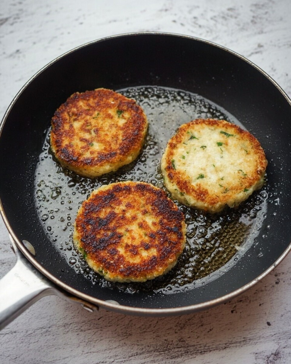 A black frying pan holds three round, thick patties cooking in oil. Two patties are golden brown and crispy on top, showing a crunchy texture with small green herbs inside. The third patty is pale and soft, without browning. The pan rests on a white marbled surface. photo taken with an iphone --ar 4:5 --v 7
