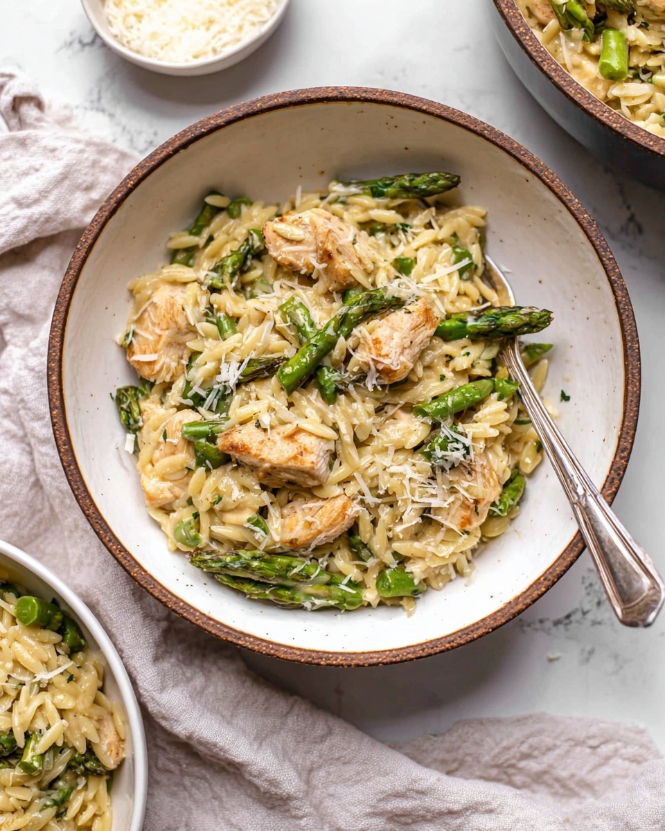 A white bowl with a brown rim holds a dish with three main layers: small pieces of light golden cooked chicken, short green asparagus pieces, and creamy light yellow orzo pasta mixed together. The orzo looks soft and slightly shiny, with the chicken and asparagus evenly spread throughout the bowl. The dish is sprinkled with white grated cheese, adding a fine texture on top. A silver fork is placed inside the bowl, resting on the food. In the background, there is a small white bowl with more grated cheese and a larger white bowl also filled with the same orzo and asparagus dish, all set on a white marbled surface with a soft white cloth beside the main bowl. Photo taken with an iphone --ar 4:5 --v 7