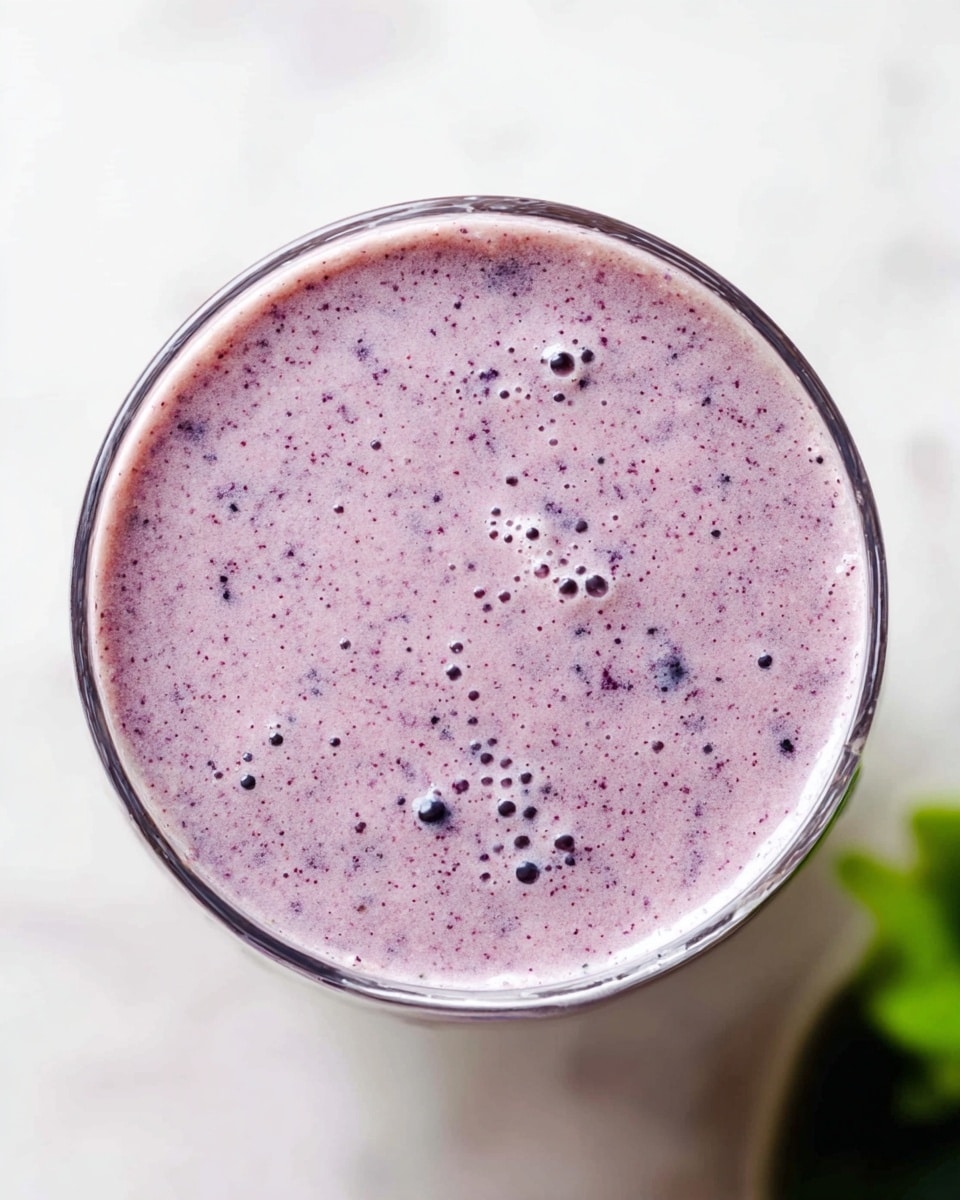 The image shows a top view of a glass filled with a creamy light purple smoothie that has small dark purple and black specks evenly spread throughout. The smoothie has a smooth texture with tiny bubbles on the surface. The clear glass is placed on a white marbled surface, and there is a small blurred green object in the background on the right side. photo taken with an iphone --ar 4:5 --v 7