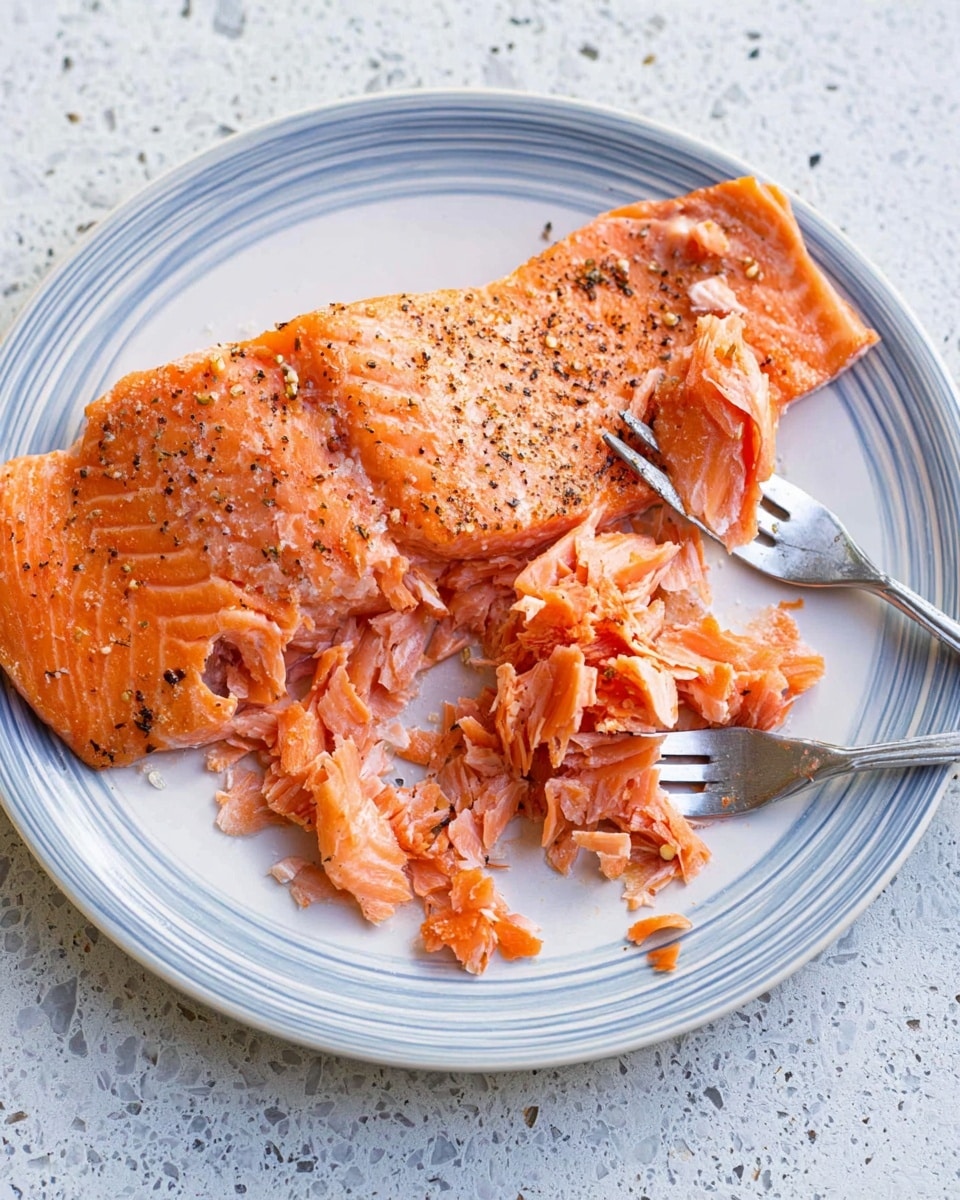 The image shows a white plate with blue rings around the edge, placed on a white marbled surface. On the plate, there is a large piece of cooked salmon with a light orange color, seasoned with black pepper and some white spots that could be salt. The salmon is partially broken into small flakes in the middle, showing its soft texture. A fork with a silver handle is placed resting on the salmon, lifting some flakes. The background surface is textured but changed to white marble. The overall look is simple and clean. photo taken with an iphone --ar 4:5 --v 7