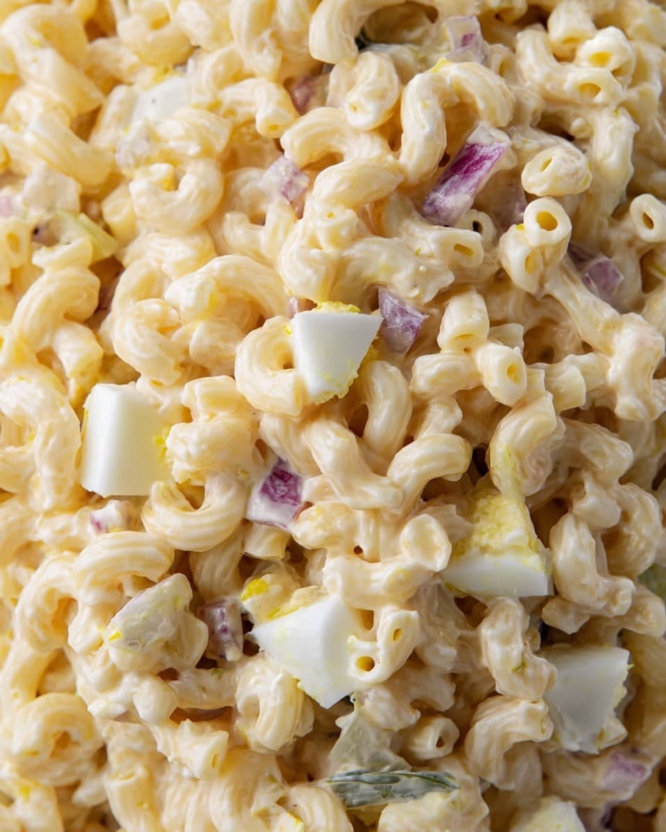 This close-up image shows a creamy macaroni salad with elbow macaroni pasta as the main layer. Mixed throughout are small pieces of chopped hard-boiled eggs, adding white and yellow contrast in the creamy, pale yellow dressing. Small bits of red onion and pickles are scattered inside, giving subtle pops of purple and green. The overall texture looks soft and smooth without any sharp edges, filling the frame fully. photo taken with an iphone --ar 4:5 --v 7