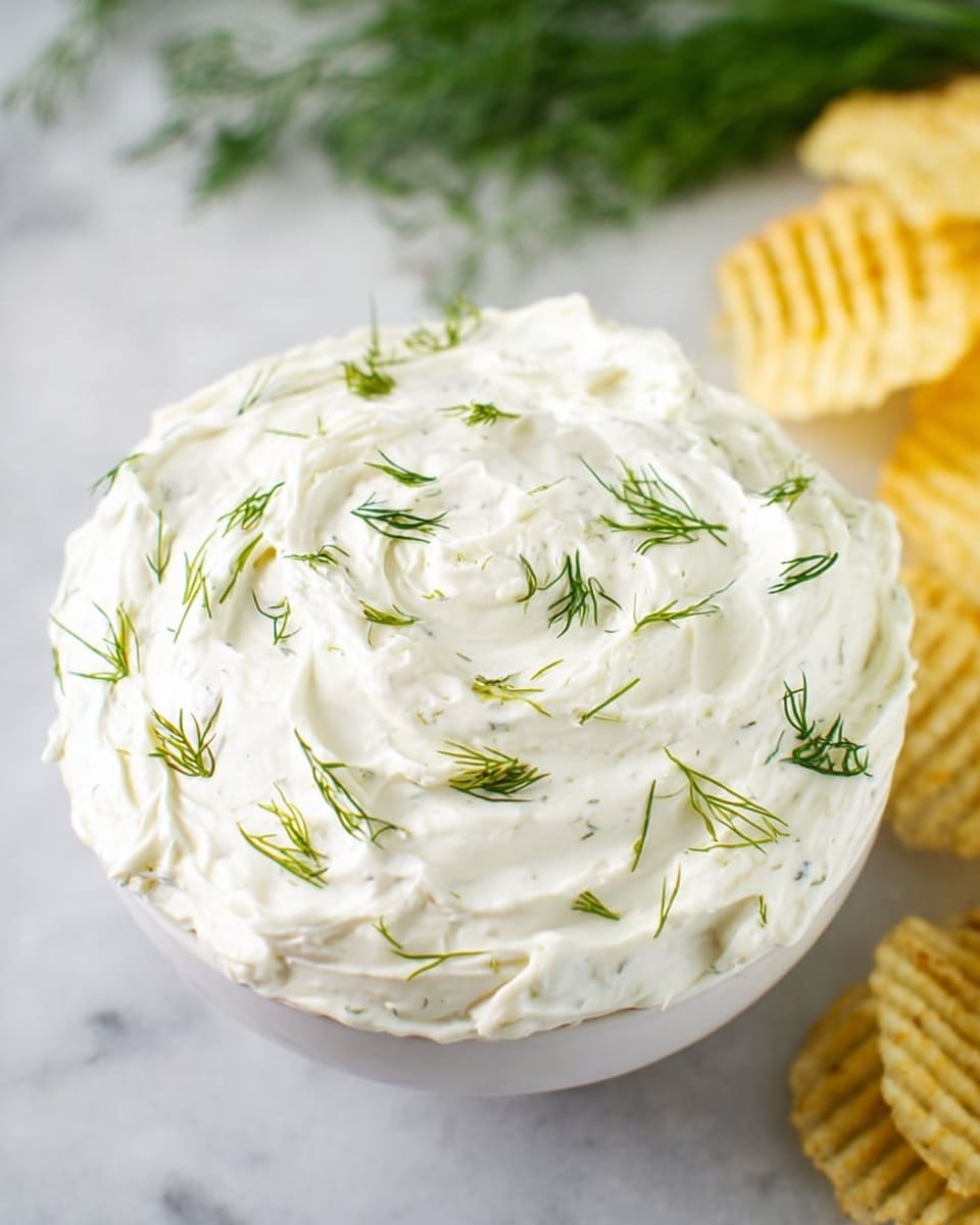 A white bowl filled with a thick, creamy white dip layered smoothly with small peaks and swirls, topped with thin, fresh green dill herb sprigs scattered unevenly on the surface. In the background on the right side, a stack of yellow ridged potato chips is slightly blurred. The bowl is placed on a white marbled surface with some blurred green herbs visible in the upper left corner. Photo taken with an iphone --ar 4:5 --v 7