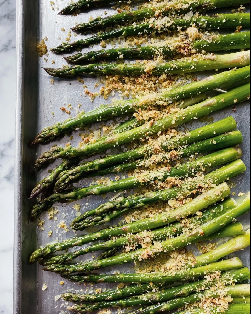 The image shows a metal tray with a single layer of green asparagus spears laid out horizontally and diagonally, topped with a sprinkling of light yellow breadcrumbs or grated cheese that adds texture and contrast. The asparagus stalks vary in shades of medium to bright green, some tips are slightly darker, and the spears have a natural woody texture visible under the topping. The tray has small scattered crumbs around the asparagus, and the background beneath the tray shows a white marbled texture. The lighting highlights the freshness and slight glossiness of the asparagus. Photo taken with an iphone --ar 4:5 --v 7