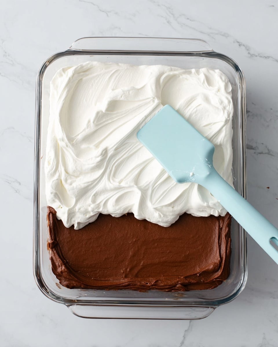 A clear glass rectangular baking dish with two layers is shown on a white marbled surface. The bottom layer is a dark brown smooth and creamy chocolate pudding spread evenly. On top of it, a thick layer of white fluffy whipped topping is being spread from the left side towards the middle, partially covering the chocolate layer. A light blue spatula rests on the right side with its edge touching the whipped topping. The whipped topping has soft swirls, giving it a light and airy texture. Photo taken with an iphone --ar 4:5 --v 7