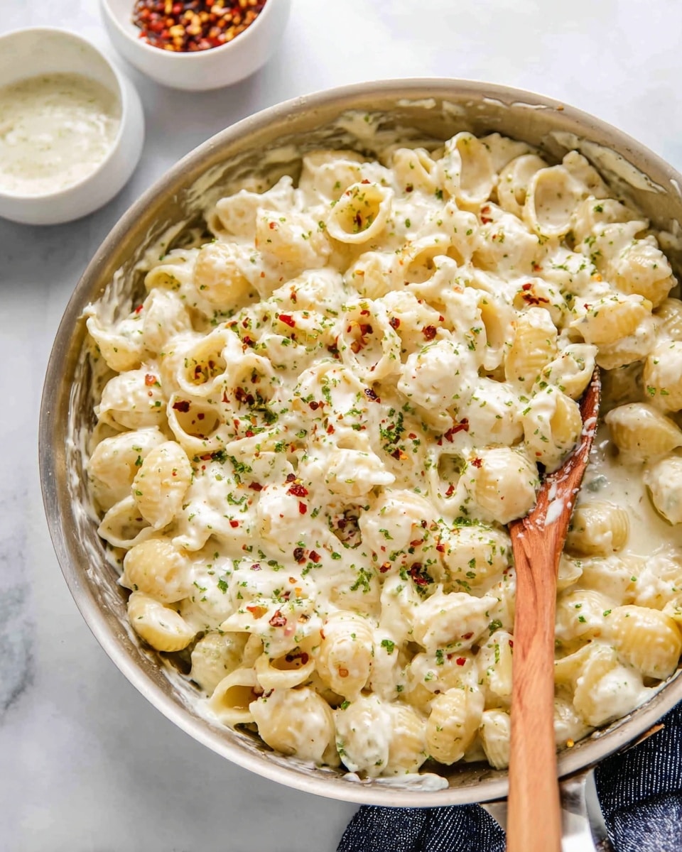 The image shows a pan full of creamy pasta shells covered in a thick, white sauce with seasoning like tiny green herbs and red chili flakes sprinkled on top. A wooden spoon is stirring the pasta near the right side of the pan. In the background, there are two small white bowls with sauces or dips, set on a white marbled surface. The pasta looks soft and creamy with a gentle texture. Photo taken with an iphone --ar 4:5 --v 7