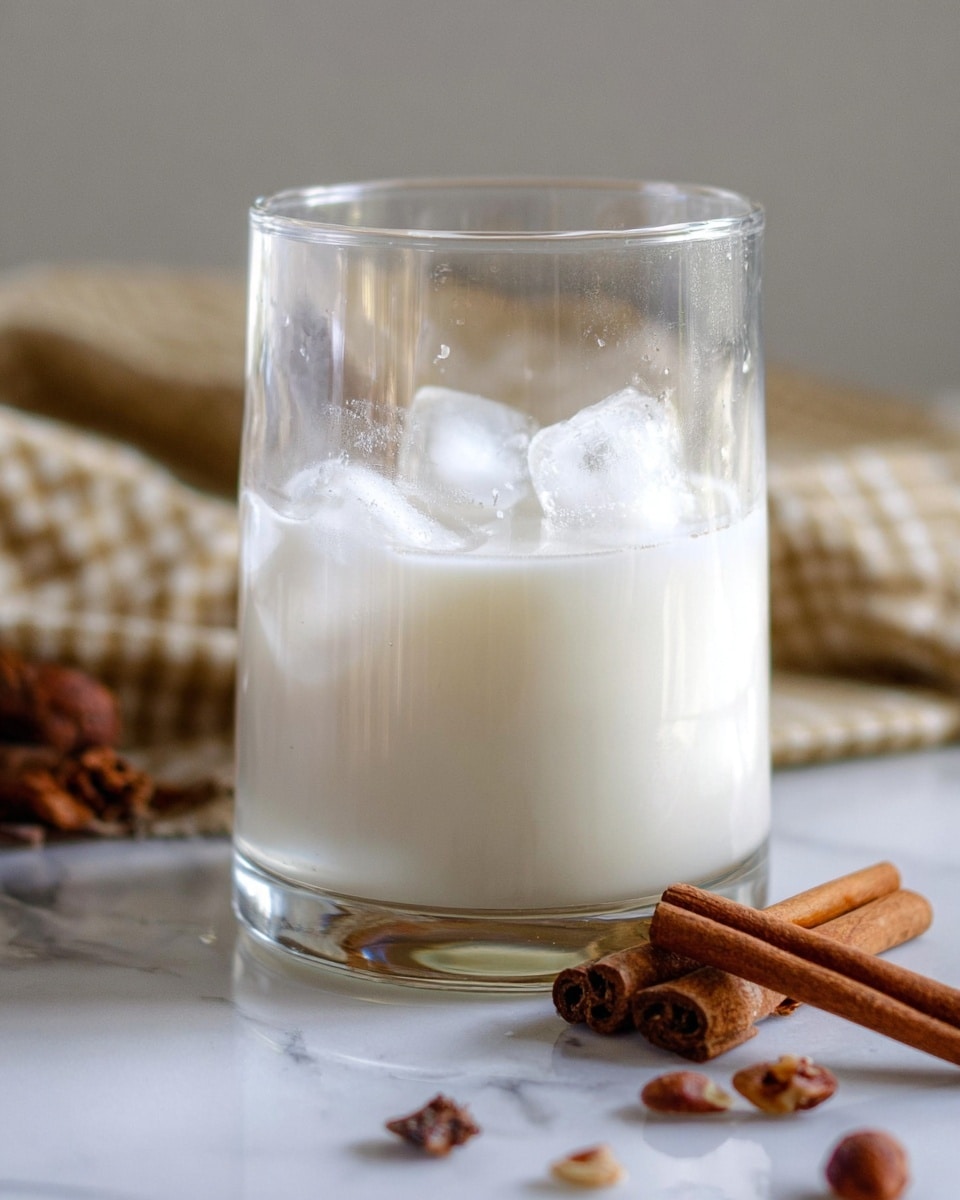 A clear glass is shown half filled with a white creamy drink with several ice cubes floating on top. The glass is placed on a white marbled surface, with two cinnamon sticks and some brown nuts scattered next to it. A folded cloth with a checkered pattern is visible in the soft blurred background. The light is natural, highlighting the smooth texture of the drink and the shiny ice cubes photo taken with an iphone --ar 4:5 --v 7