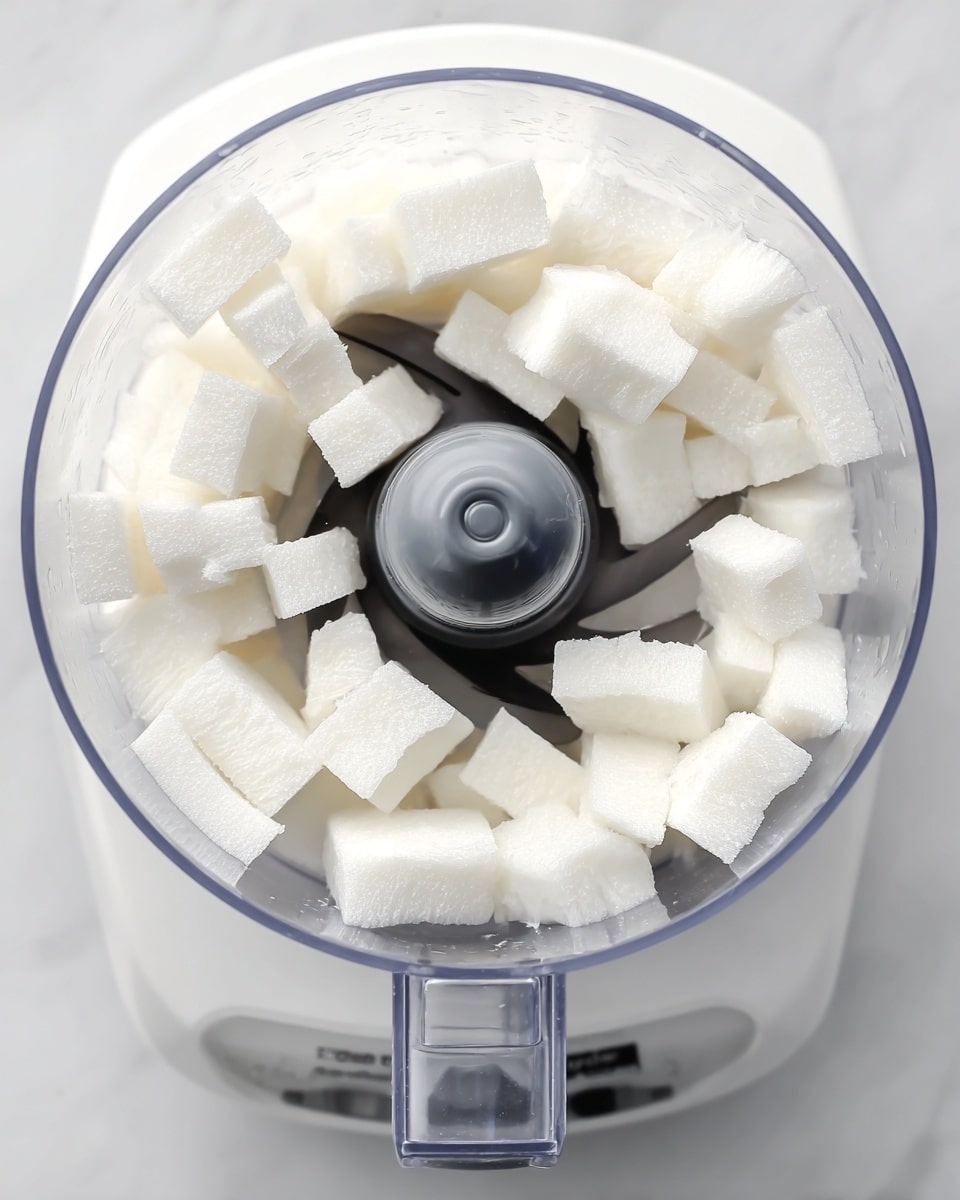 A clear food processor bowl filled with small, thick white rectangular chunks layered evenly around the central black and grey blade. The bright white pieces have a smooth and firm texture, sitting on a white marbled surface visible through the transparent bowl. The food processor is white with clear plastic parts, and the image is taken from above, showing the clean and simple setup. photo taken with an iphone --ar 4:5 --v 7