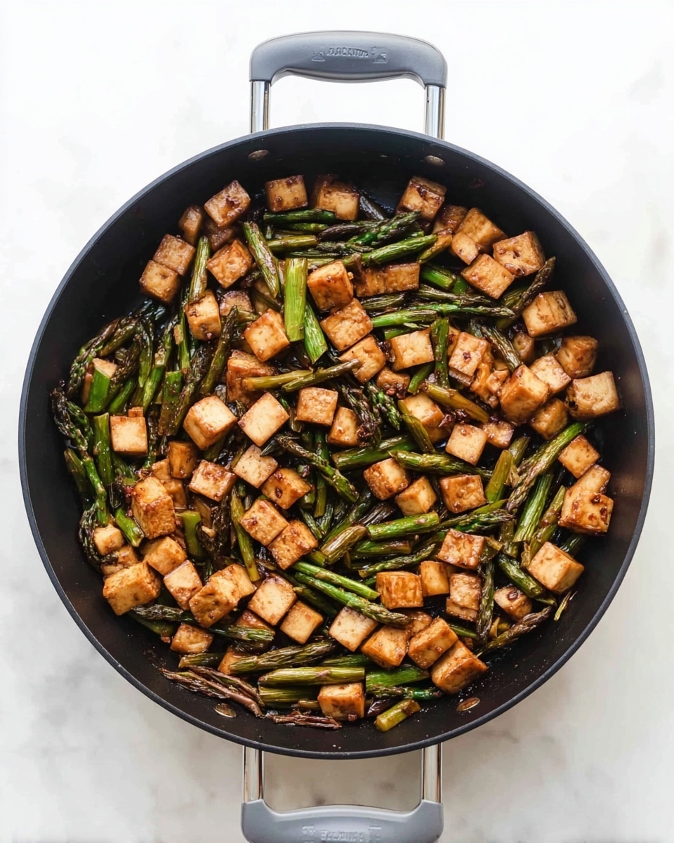 A black pan filled with cooked small orange-brown cubes of tofu and long green asparagus pieces slightly charred, mixed evenly throughout. The tofu cubes have a firm texture with some browning on edges, and the asparagus spears are slender with a tender look. The pan has a dark inside surface and two handles with gray grips, placed on a white marbled textured surface photo taken with an iphone --ar 4:5 --v 7