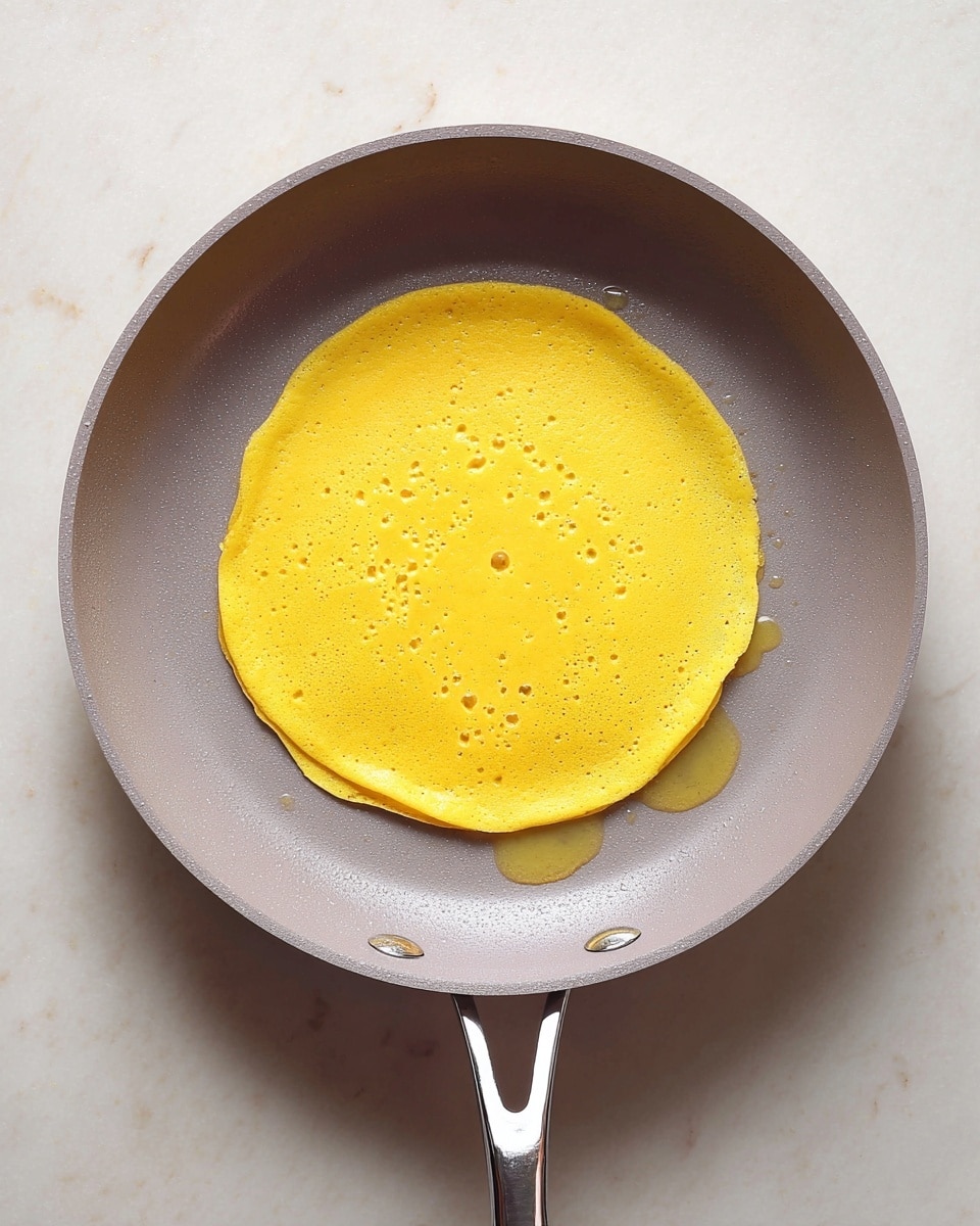 A top-down view of a single layer of a bright yellow pancake cooking in a gray non-stick frying pan with a silver handle. The pancake has a smooth texture with small bubbles forming on the surface and a slightly uneven round shape with a gently scalloped edge. Drops of oil are visible around the pancake in the pan. The frying pan sits on a white marbled surface. photo taken with an iphone --ar 4:5 --v 7