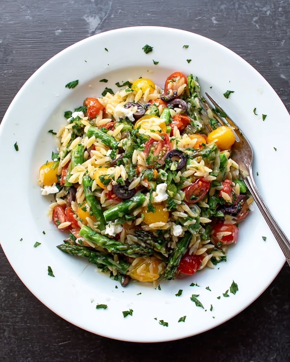 A white plate holds a colorful orzo salad with three main layers. The bottom layer is made of light beige orzo pasta mixed with green asparagus pieces positioned throughout. The middle layer features bright yellow and red cherry tomatoes, halved, scattered evenly. The top layer is sprinkled with fresh green chopped herbs, small white cheese chunks, and black olive slices. A fork rests on the right side of the plate, its prongs inside the salad, with scattered herbs around the plate edge, all set on a white marbled surface photo taken with an iphone --ar 4:5 --v 7