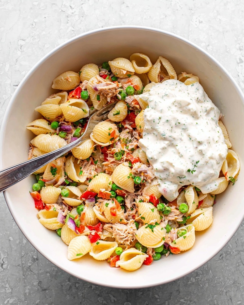 A white bowl filled with a shell pasta salad that has a mix of ingredients evenly spread. The base layer consists of light yellow shell pasta, scattered with small green peas, red bell pepper pieces, celery bits, shredded light brown tuna, and small chunks of red onion. On the right side of the bowl, there is a thick dollop of white creamy dressing with green herb specks on top of the pasta mixture. A shiny metal fork is placed inside the bowl on the left side, resting among the ingredients, and the bowl is set on a white marbled textured surface. Photo taken with an iphone --ar 4:5 --v 7