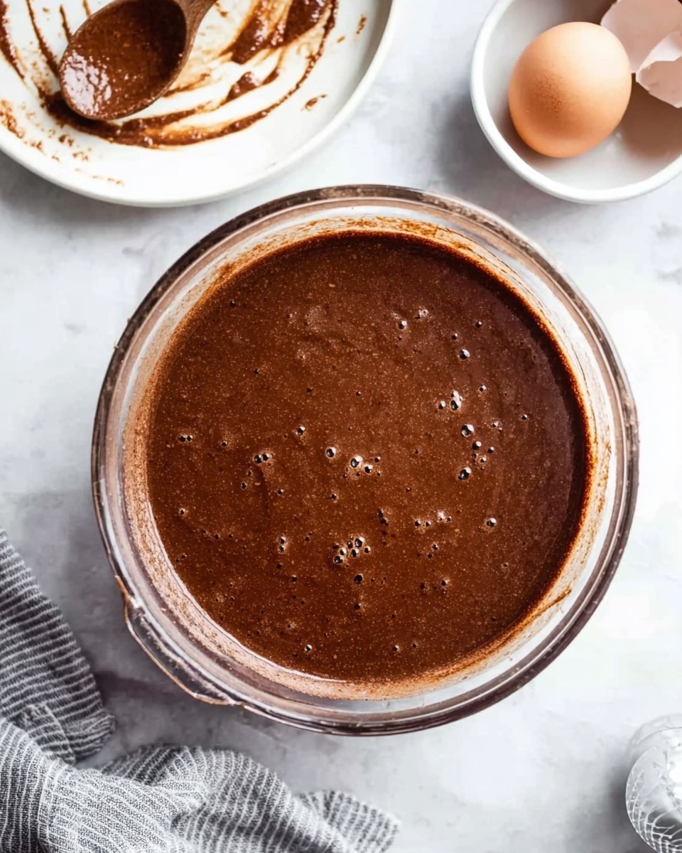 A clear glass mixing bowl filled with smooth, dark brown chocolate batter with a few small bubbles on the surface is placed on a white marbled texture. Behind the bowl is a white plate with some smears of chocolate and a wooden spoon resting on it, lightly coated with chocolate. To the right edge of the image, there is a small round white bowl holding two cracked eggs with visible eggshell halves. A gray and white striped cloth is partially visible at the bottom right corner. Photo taken with an iphone --ar 4:5 --v 7