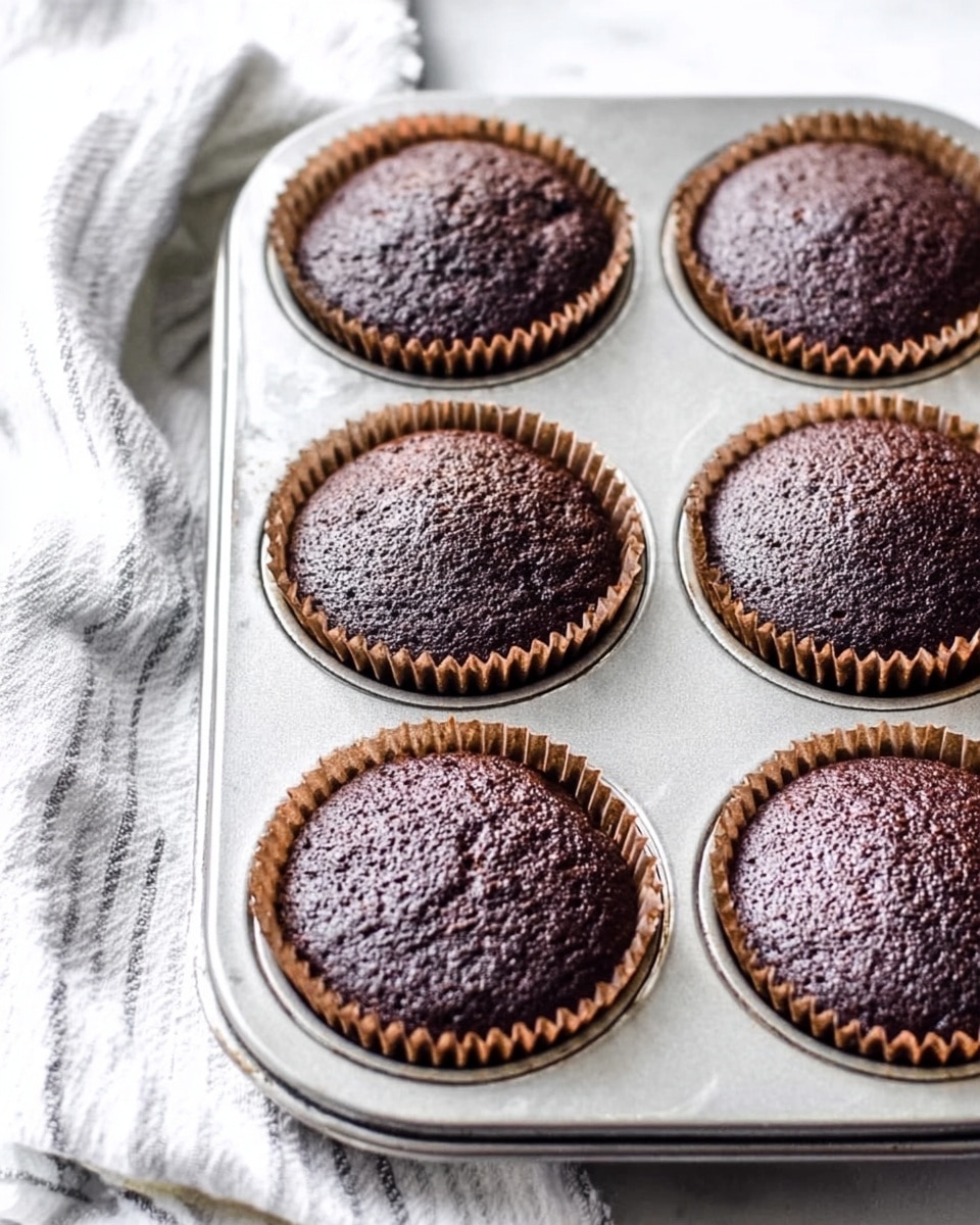The image shows six dark brown chocolate cupcakes in a silver metal cupcake tray, each in a brown paper liner with ridged edges. The cupcakes have a smooth, slightly rough textured top with some small cracks and dimples. The tray is placed on a white marbled surface next to a white cloth with thin gray stripes. The overall scene is bright with soft natural light, highlighting the rich color and texture of the cupcakes. photo taken with an iphone --ar 4:5 --v 7