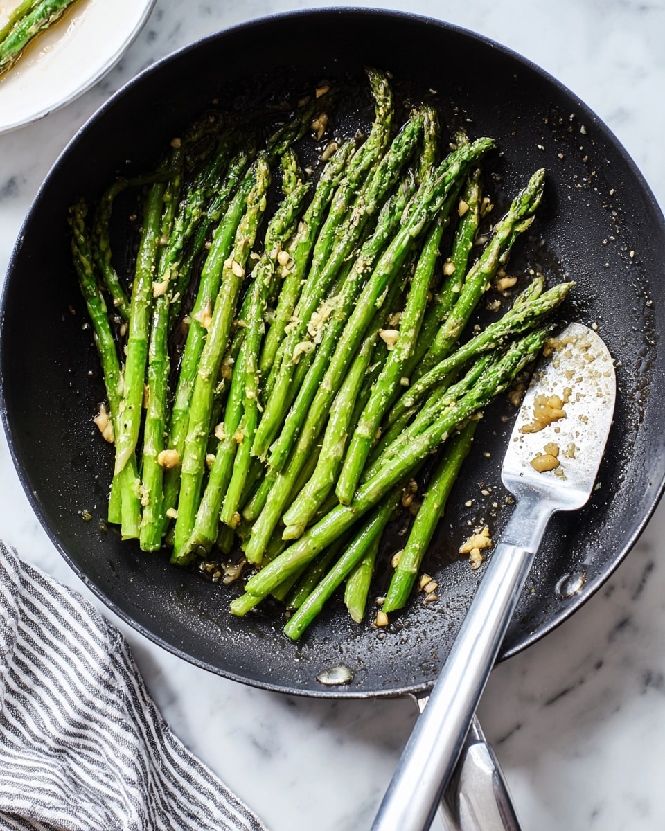 A black frying pan filled with about two dozen bright green asparagus spears, cooked and slightly charred, arranged in a loose cluster mostly on the left side of the pan. Small bits of chopped garlic are scattered on the asparagus and around the pan. A gray spatula with a silver handle rests inside the pan on the right side. The pan sits on a white marbled surface with a striped cloth towel partially visible at the bottom left corner. Photo taken with an iphone --ar 4:5 --v 7