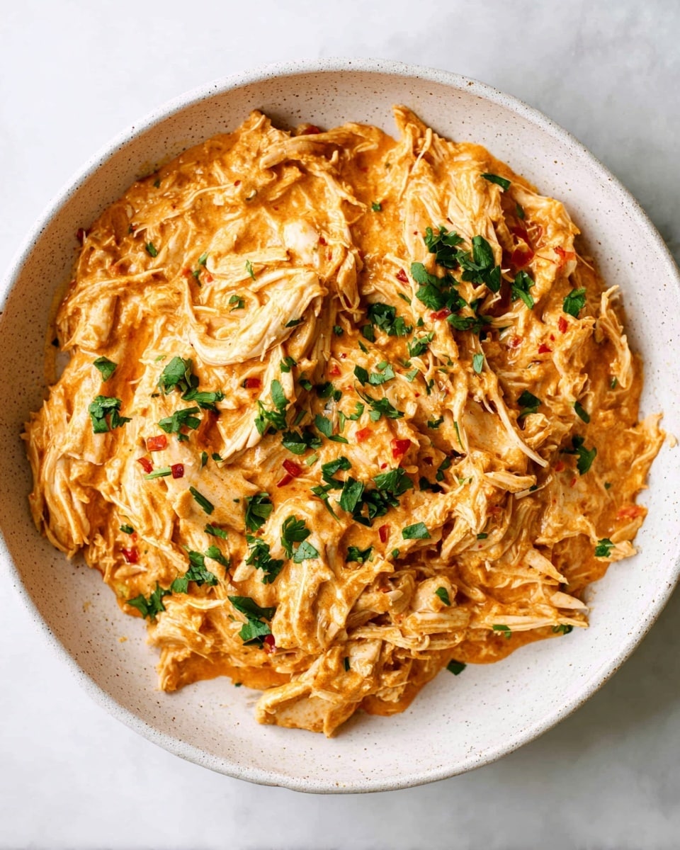 The image shows a bowl filled with shredded chicken coated in a creamy orange sauce that looks rich and smooth. The chicken pieces appear soft and fibrous, mixed well with the thick sauce that includes small red and green specks. On top, bright green chopped herbs add a fresh touch of color. The bowl is white with a slightly speckled texture around the edges, and the whole scene sits on a white marbled surface, giving it a clean and bright look. photo taken with an iphone --ar 4:5 --v 7