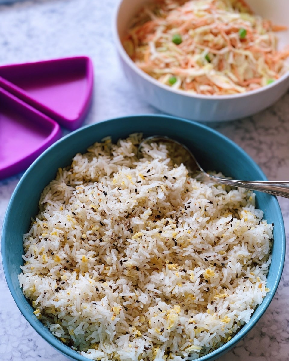 The image shows a blue bowl filled with cooked white rice mixed with small black, yellow, and light brown seasoning pieces, giving the rice a speckled texture. The rice is fluffy and loose, with a spoon partially stuck into the right side of the bowl. In the background, there is a white bowl containing a creamy mix of shredded pale orange and white ingredients, likely a salad or coleslaw, with small green bits scattered throughout. Next to the bowls, there are two purple triangular kitchen tools placed on a white marbled surface. The overall scene has a soft, natural light quality. photo taken with an iphone --ar 4:5 --v 7