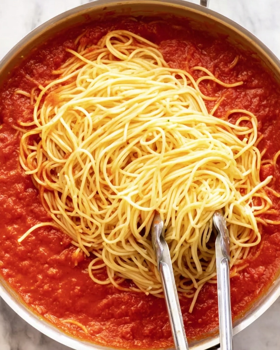 In a close-up view, a cooking pan filled with a thick, bright red tomato sauce covers the bottom layer. On top of the sauce, there is a large pile of light yellow cooked spaghetti noodles with a soft texture, loosely spread over the sauce. Two silver metal tongs rest over the noodles, positioned diagonally with one end touching the sauce and the other end above the pile. The pan is set on a white marbled surface. photo taken with an iphone --ar 4:5 --v 7