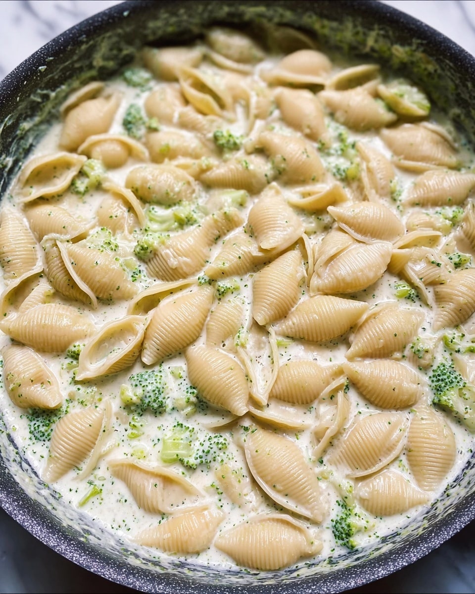 The image shows a close-up of pasta shells cooking in a creamy sauce with small green bits of broccoli mixed in. The pasta shells are light beige, smooth with ridges on the surface, and are evenly spread throughout the creamy white sauce that has a slightly foamy texture. Small pieces of bright green broccoli are visible, adding a fresh contrast of color. The dish is contained inside a dark pan, set against a white marbled surface. The overall look is gooey and soft with a mix of smooth and chunky textures. photo taken with an iphone --ar 4:5 --v 7