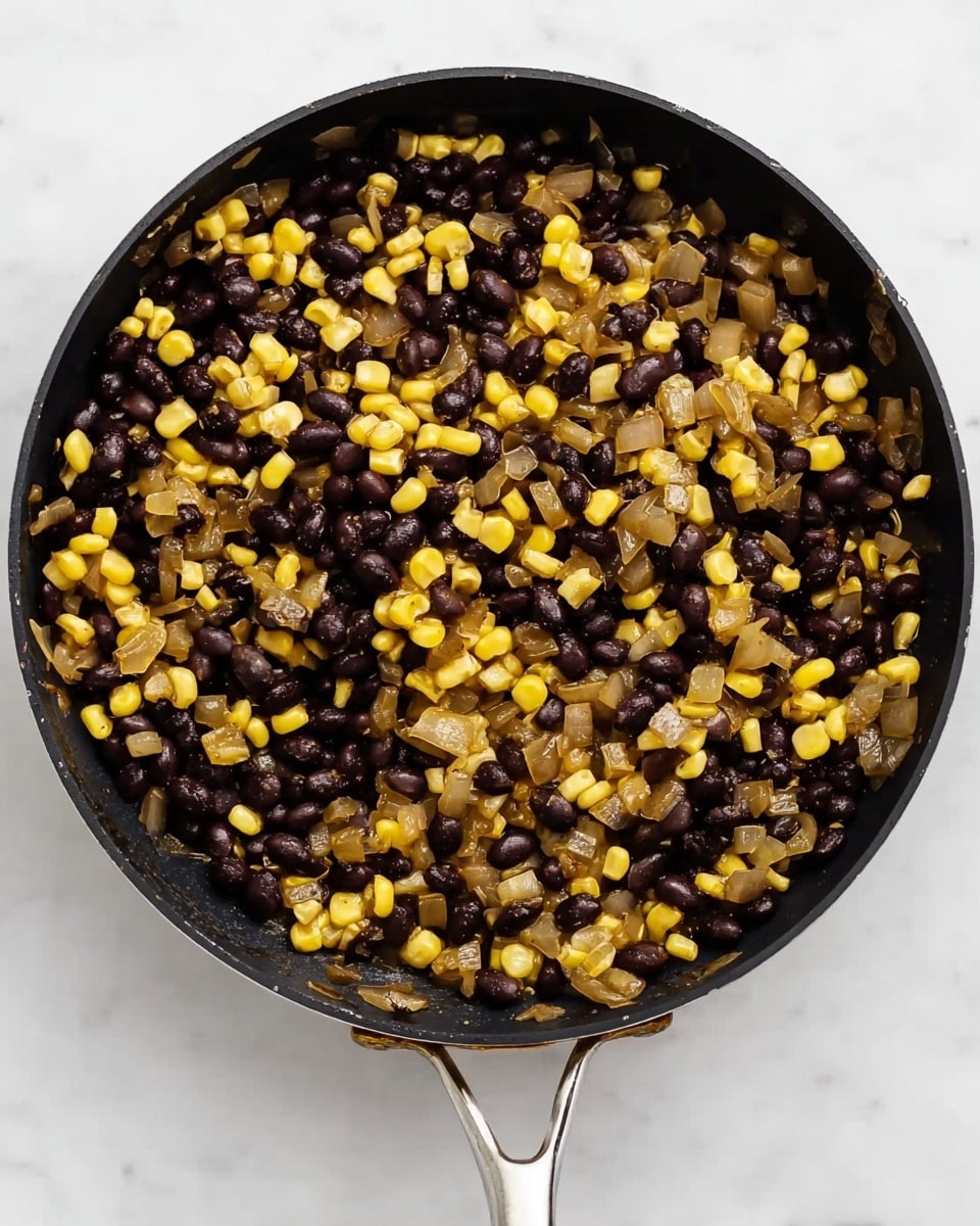 A black pan filled with a mix of black beans, bright yellow corn, and translucent cooked onion pieces, all lightly seasoned and cooked together evenly. The black beans and yellow corn create a colorful contrast, with the onions adding a slightly glossy texture throughout. The pan sits on a white marbled surface, and its silver handle is visible at the bottom of the image. photo taken with an iphone --ar 4:5 --v 7