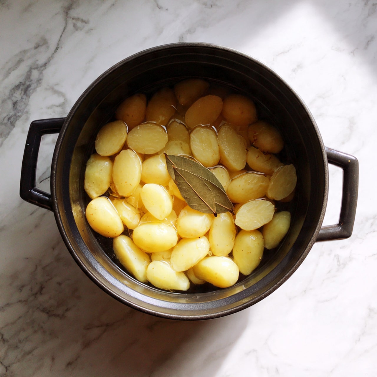A black pot filled with water and many light yellow potato pieces evenly spread inside, with one light brown bay leaf placed on top near the center. The pot sits on a white marbled surface. Photo taken with an iphone --ar 4:5 --v 7