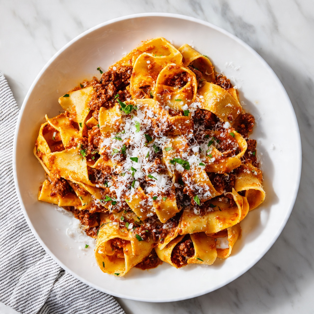 A white round plate is filled with a generous serving of wide, flat pasta ribbons coated in a rich, deep orange-red sauce mixed with chunks of dark brown meat. The pasta strands twist and curl naturally, showing their slightly rough texture. The dish is sprinkled with grated white cheese and small green herb leaves scattered all over the top, adding fresh color contrast. The plate rests on a white marbled surface with a striped cloth visible at the bottom left corner. Photo taken with an iphone --ar 4:5 --v 7