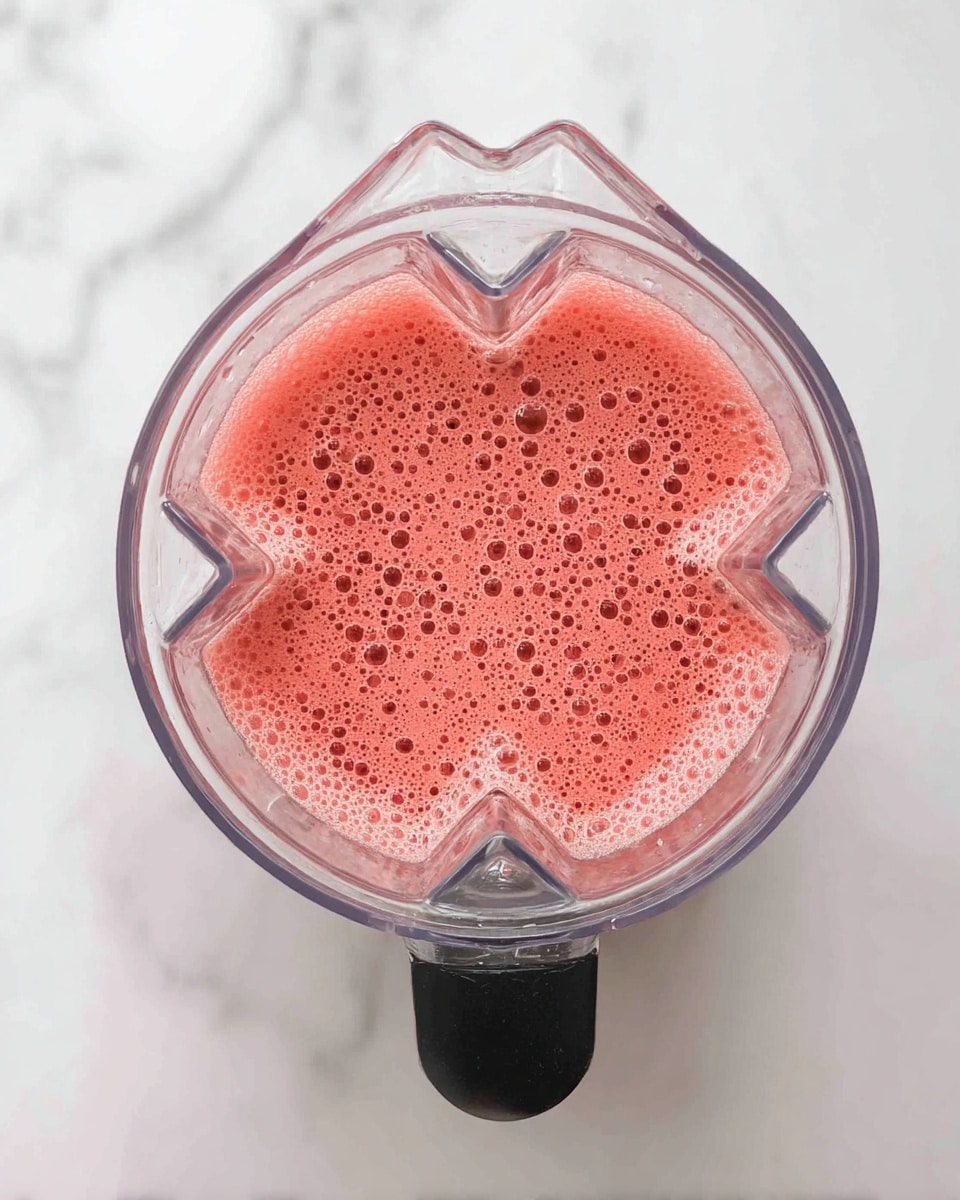 A clear blender jar is shown from above, filled with a pinkish-red liquid that has many small bubbles on its smooth surface. The blender jar has a clear, faceted texture with a black handle at the bottom center. The whole scene is set on a white marbled surface. photo taken with an iphone --ar 4:5 --v 7