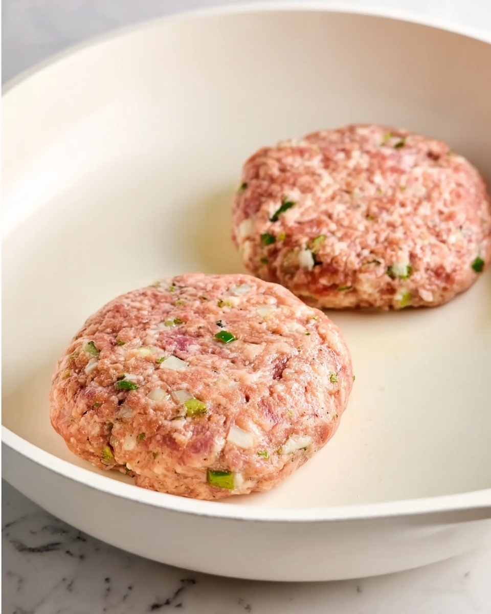 Two round uncooked patties are in a white frying pan with a smooth surface. The patties have a light pink color with white and green bits visible throughout, showing textures of chopped ingredients mixed inside. The inside of the pan is clean and shiny with a simple light tone. The background shows a white marbled texture. Photo taken with an iphone --ar 4:5 --v 7