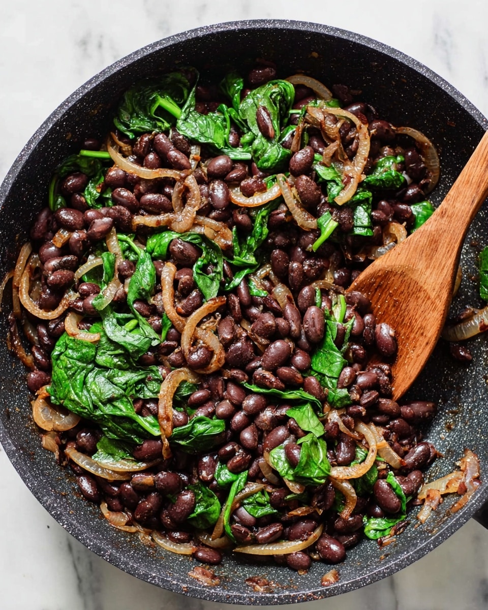 A close-up of a black non-stick pan filled with a cooked mix of three main layers: at the bottom, dark brown to black cooked beans form the bulk with a smooth, round shape; mixed on top throughout are thin, golden-brown cooked onion rings that look slightly soft and shiny; scattered in between are bright green wilted spinach leaves, soft in texture but still vibrant. A wooden spoon rests inside the pan on the right side, partly covered by the beans and greens. The pan is placed on a white marbled surface. Photo taken with an iphone --ar 4:5 --v 7