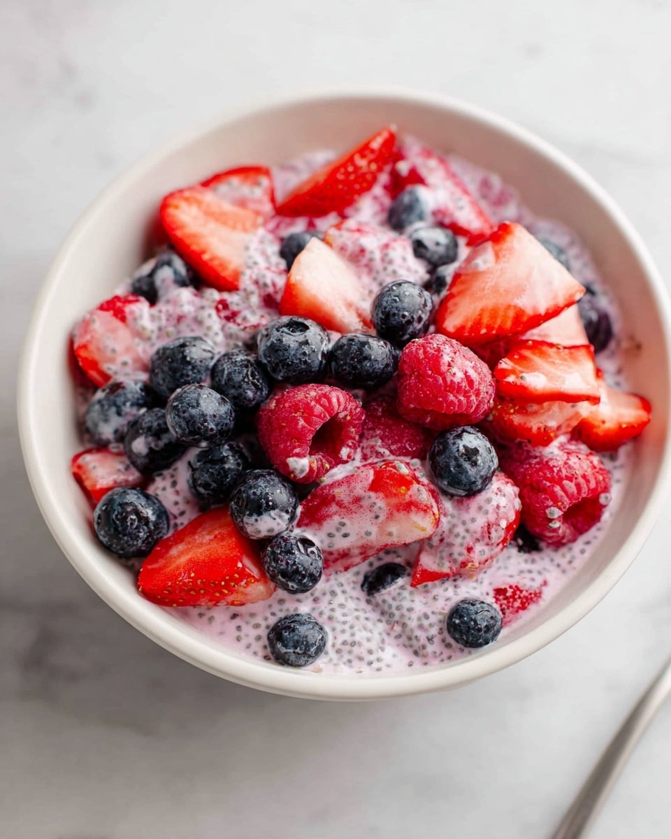 A white bowl filled with a creamy mixture covered with three kinds of berries on a white marbled surface; the base layer is light pink cream mixed with tiny black seeds giving a smooth yet slightly textured look; on top of this layer are whole dark blue blueberries scattered all over; mixed in are red raspberries and sliced strawberries that add bright red and light pink colors, some strawberry slices showing their inner white flesh; the berries sit both on top and partially covered by the cream, creating a rich, colorful, and fresh look; photo taken with an iphone --ar 4:5 --v 7