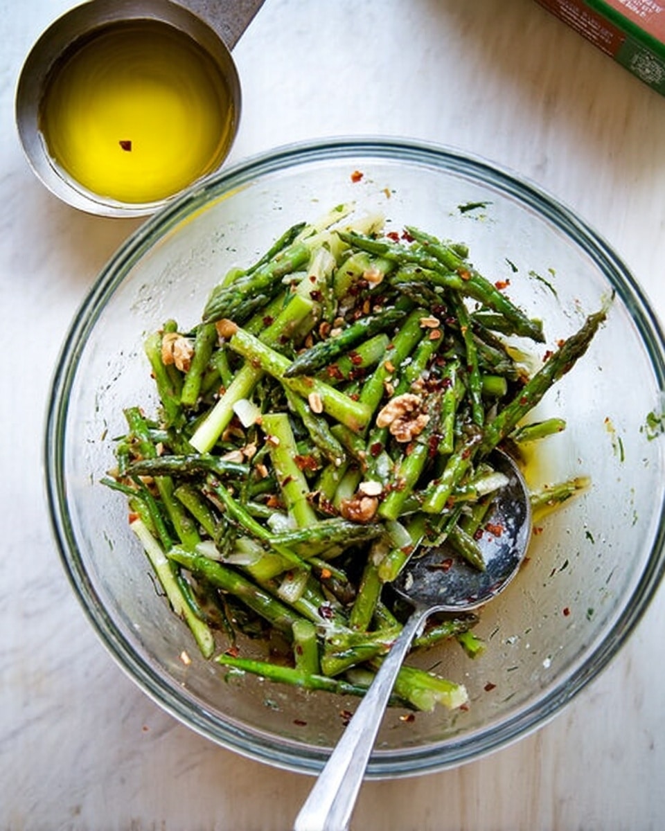 A clear glass bowl filled with a mix of thin green asparagus slices, some slightly charred, mixed with small bits of nuts and red chili flakes, all coated lightly with a dressing that gives a glossy texture. A silver spoon rests inside the bowl on the right side, and to the left, there is a metal measuring spoon holding yellow olive oil. The bowl is placed on a white marbled surface, and a box of olive oil is partially visible on the upper right corner. Photo taken with an iphone --ar 4:5 --v 7