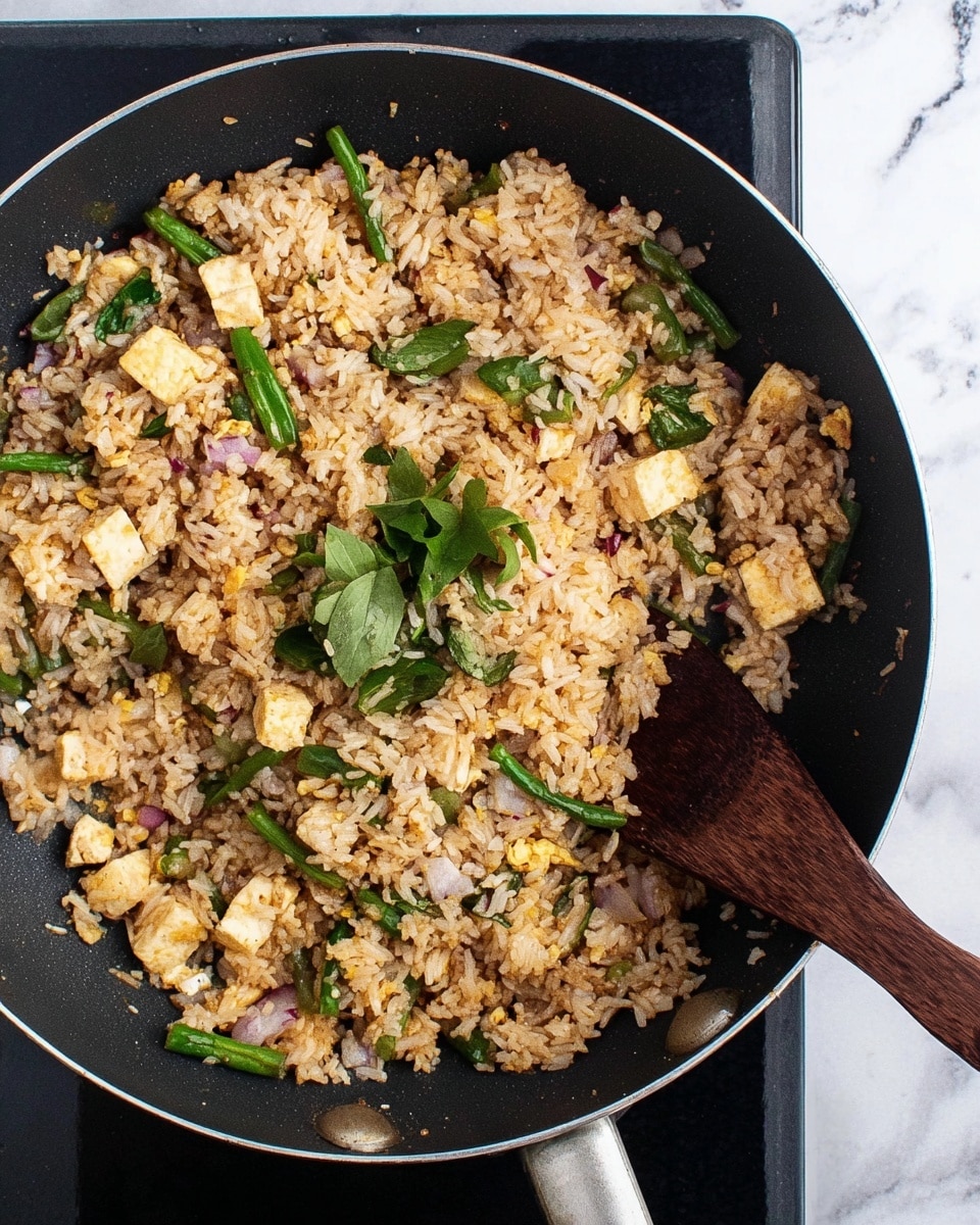 The image shows a black frying pan filled with mixed fried rice. The rice is light brown and mixed with small cubes of pale yellow vegetable pieces, green beans cut into short pieces, and some green leafy herbs scattered on top. There are also small bits of white tofu and red onion pieces mixed throughout the rice. A dark wooden spatula rests inside the pan, partially stirring the rice. The pan sits on a black cooking stove with a white marbled surface beneath it. photo taken with an iphone --ar 4:5 --v 7