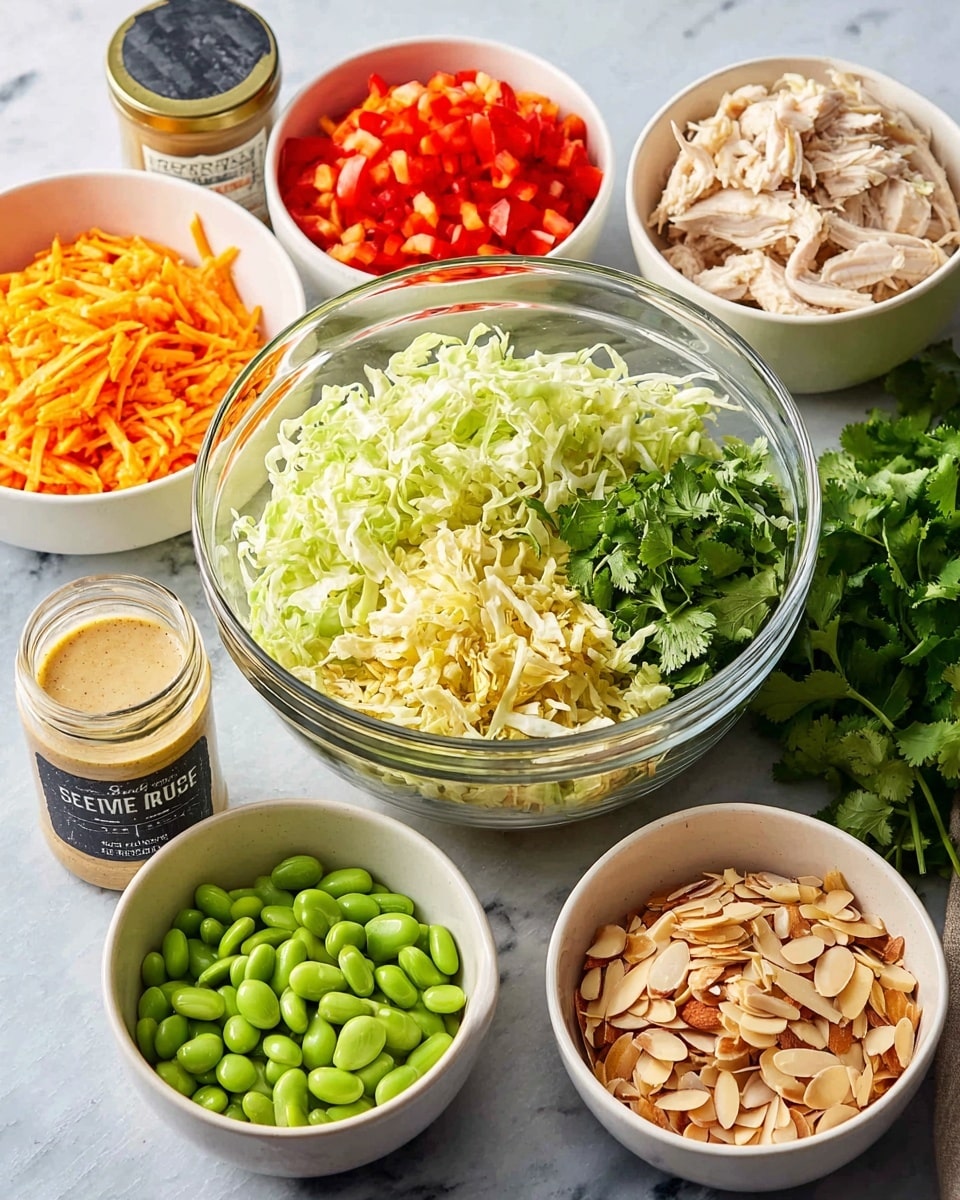The image shows six bowls arranged on a white marbled surface, each holding different fresh ingredients for a salad. In the large clear glass bowl at the center, there are two layers of leafy vegetables: one side is pale yellow, finely shredded cabbage, and the other side is bright green, chopped lettuce. Behind the big bowl are two white bowls; one on the left contains shredded bright orange carrots and red diced bell peppers, while the one on the right has light beige shredded cooked chicken. In front of the big bowl and slightly to the left is a white bowl filled with smooth green edamame beans. In front of the big bowl and slightly to the right is a white bowl with toasted almond slices, light brown with some darker edges. Between the edamame and almonds is a small glass jar with a creamy beige sesame seed dressing. To the left side, there is a jar labeled