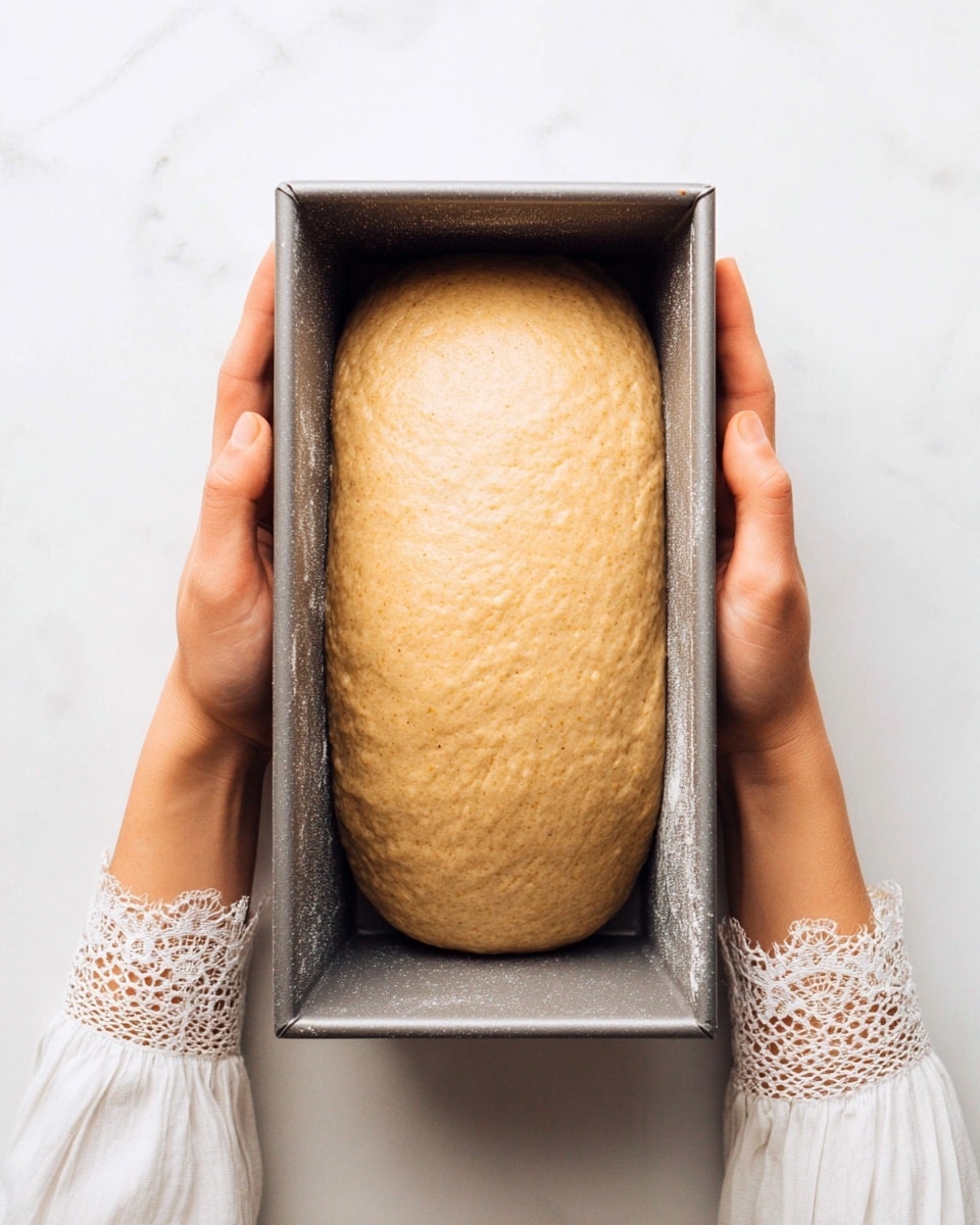 A gray metal loaf pan holds a single smooth ball of light brown dough with a soft texture, filling the pan almost to the edges. The pan is held by both sides with a woman's hands wearing a white long-sleeve top with lace cuffs. The background is a white marbled texture. photo taken with an iphone --ar 4:5 --v 7