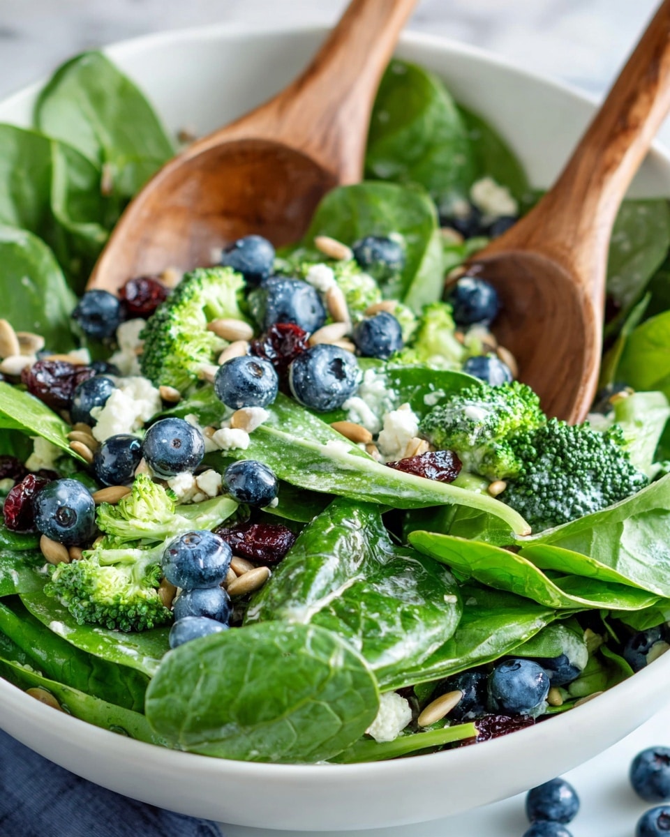 A close-up of a fresh salad in a white bowl filled with bright green spinach leaves and small broccoli pieces as the bottom layer. On top, there are scattered blueberries, dried cranberries, and small chunks of white cheese. Light brown sunflower seeds are sprinkled throughout the mix. Two wooden salad spoons are partially placed in the bowl, mixing the ingredients together. The bowl is on a white marbled surface with a few blueberries beside it. photo taken with an iphone --ar 4:5 --v 7
